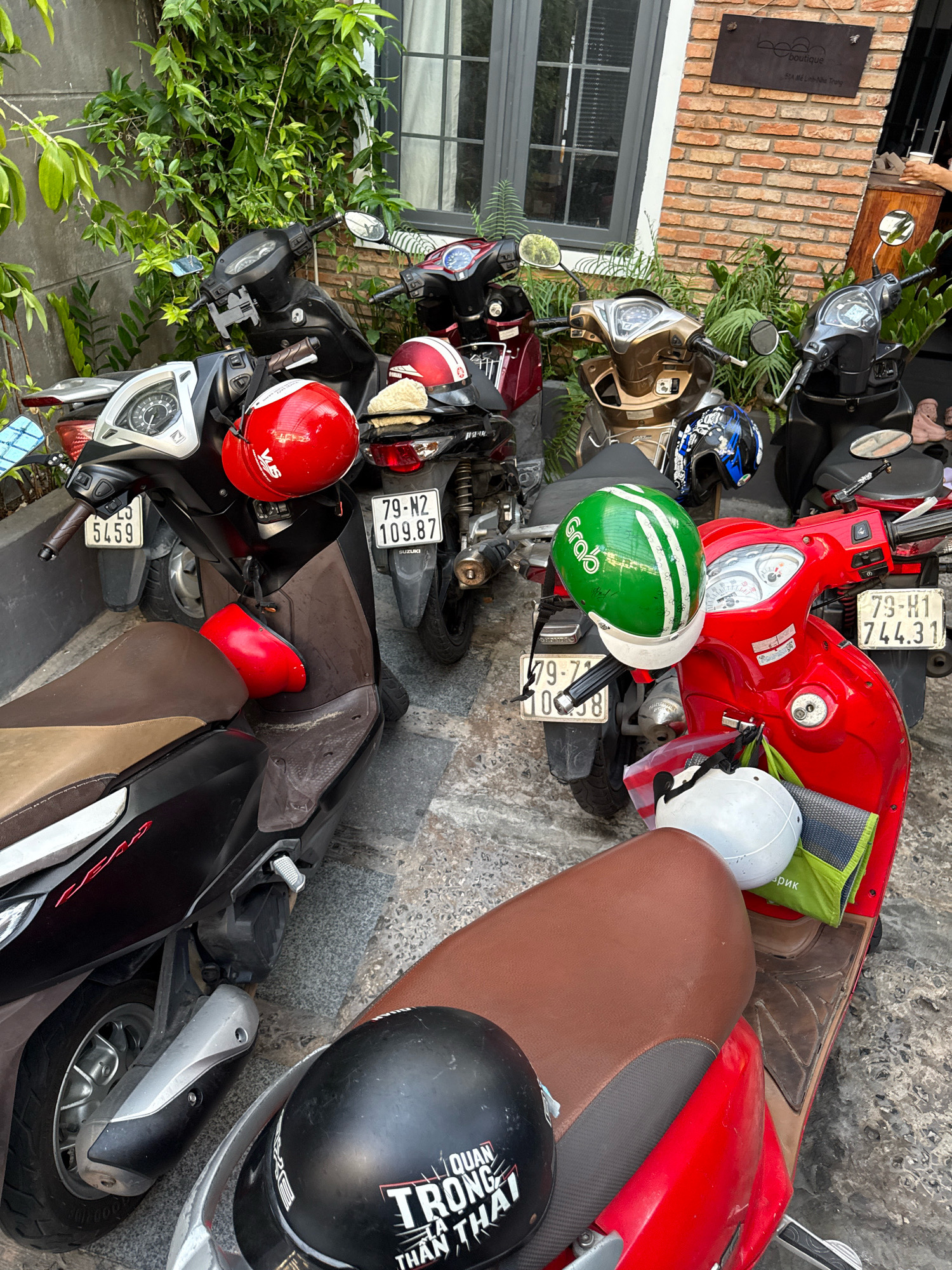 The image depicts a street scene in Nha Trang, Vietnam, likely in front of a building with a brick facade and a sign that reads "Boutique". Numerous motorcycles are parked closely together on a paved surface. Several helmets are visible, including a green Grab helmet, two red helmets, a blue and black helmet, and a black helmet with white lettering. The motorcycles appear to be in operational condition, with license plates visible on some of them. Green foliage and plants are growing around and behind the motorcycles, indicating an outdoor setting. The overall impression is one of a common sight in urban areas of Vietnam where motorcycles are a primary mode of transportation.