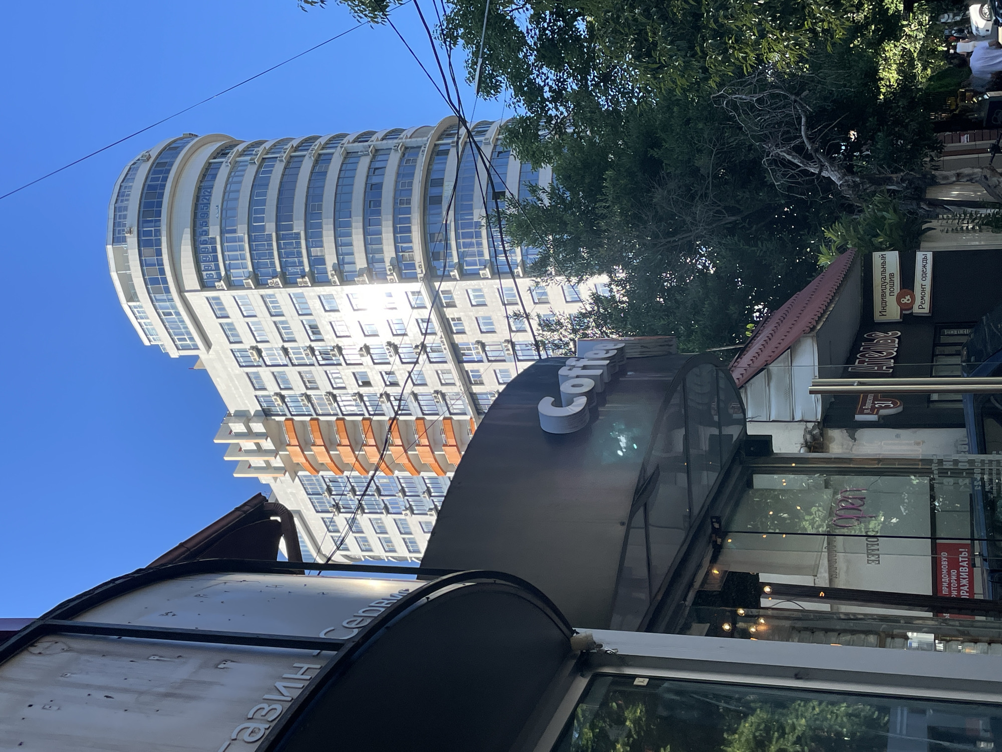 The image shows a street scene in Sochi, Russia, on a clear day. In the foreground, a curved, dark awning covers a storefront with a glass facade. The awning has raised white lettering that appears to read "магазин" and "Сервис". Below the awning, the glass entrance to a business is visible, with reflections of trees and lights. The word "open" is visible in pink lettering on the glass door. A sign in Russian that translates to "territory" and "protected" is partially visible. Next to this, another sign reads "Individual tailoring" and "Clothing repair".
Behind the storefront, a tall, modern residential building with a curved facade dominates the background. The building is light-colored with numerous windows and balconies, some of which have orange railings. The upper floors of the building are rounded. Several air conditioning units are visible on the exterior walls. Overhead power lines stretch across the frame.
To the right of the residential building, dense green trees are visible, some with bare, twisted branches. The sky is a clear, bright blue. A few people and a car are visible on the sidewalk to the far right of the frame. A red-tiled roof of a smaller building is visible in the lower right quadrant. The overall impression is an urban street scene with a mix of older and newer architecture.