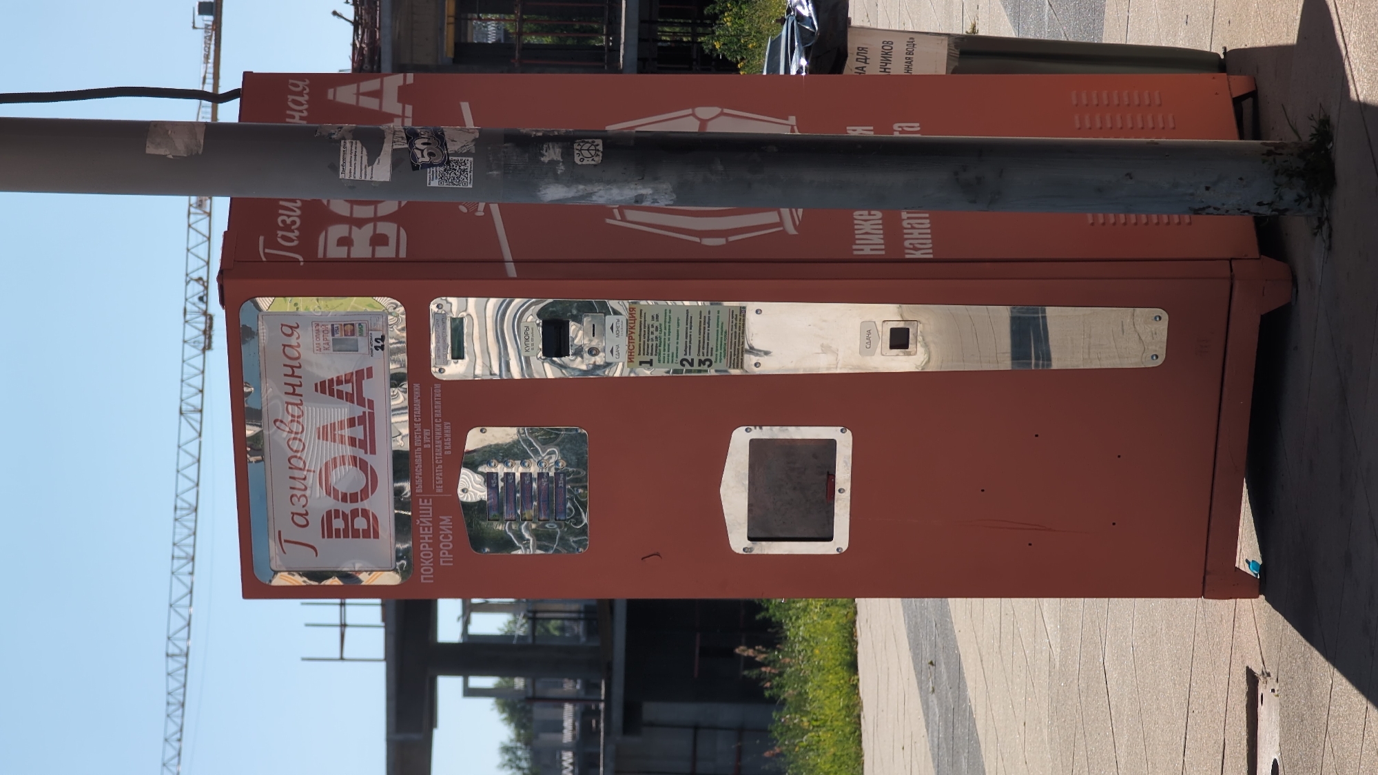 The image shows a red vending machine for carbonated water on a paved sidewalk in Bor, Russia. The machine is tall and rectangular, with a metal pole in front of it. The pole is covered in stickers and tape. The vending machine has a large display on the front that reads "Газированная ВОДА" (Carbonated Water) in Russian. Below this, there are instructions in Russian on how to use the machine, including options for payment by card or coin. There are also slots for inserting money and receiving change. In the background, a construction crane is visible against a bright blue sky, suggesting it is daytime with clear weather. The machine appears to be in a public area, possibly a park or street. The overall scene depicts a typical urban environment with functional public amenities.