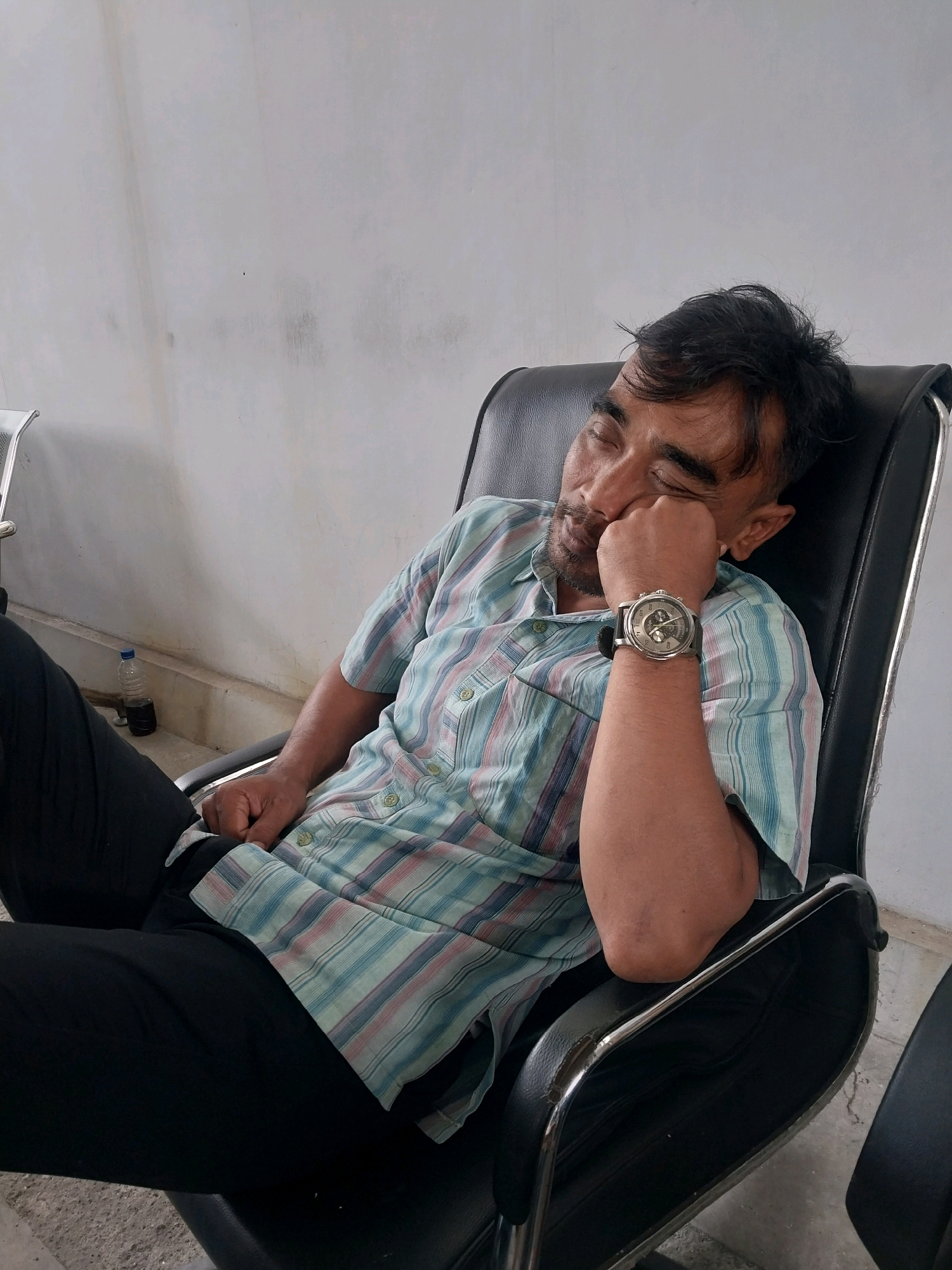A man is asleep in a black office chair with chrome accents. He is wearing a light blue and pink striped button-down shirt and black pants. His right arm is resting on his face, and he is wearing a silver wristwatch on his left wrist. A plastic water bottle with dark liquid is visible on the floor to the left of the chair. The background consists of a plain, light-colored wall. The scene appears to be indoors, possibly in an office waiting area or similar public space in Sleman, Indonesia.