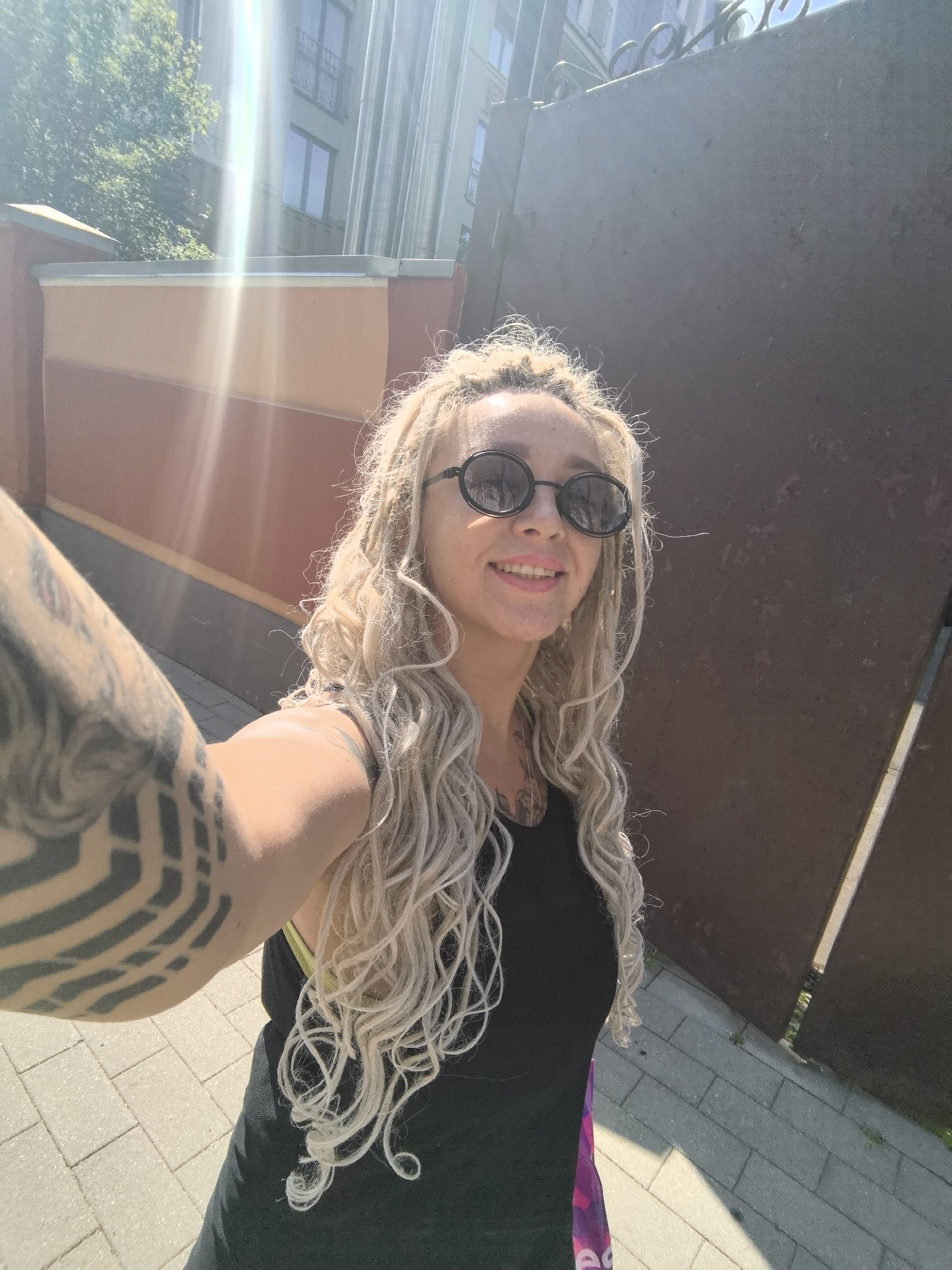 A woman with blonde dreadlocks and extensive arm tattoos is taking a selfie outdoors on a sunny day. She is wearing round, dark sunglasses and a black tank top, with a hint of a bright purple and pink patterned fabric visible at the bottom of her attire, possibly a bag or part of her clothing. The woman is smiling at the camera, appearing happy and relaxed.
The background suggests an urban or semi-urban setting. To her left, there is a building with multiple floors and windows, partially obscured by sunlight creating lens flares. A textured wall with reddish-brown and tan sections runs horizontally in front of the building. To her right, a large, dark, textured metal surface dominates the background, possibly a fence or a wall. The ground is paved with interlocking light-colored bricks, and some greenery is visible peeking through the gaps between the bricks. The strong sunlight indicates it is daytime, likely midday given the angle of the shadows. The overall atmosphere is bright and cheerful. No visible text is present in the image.