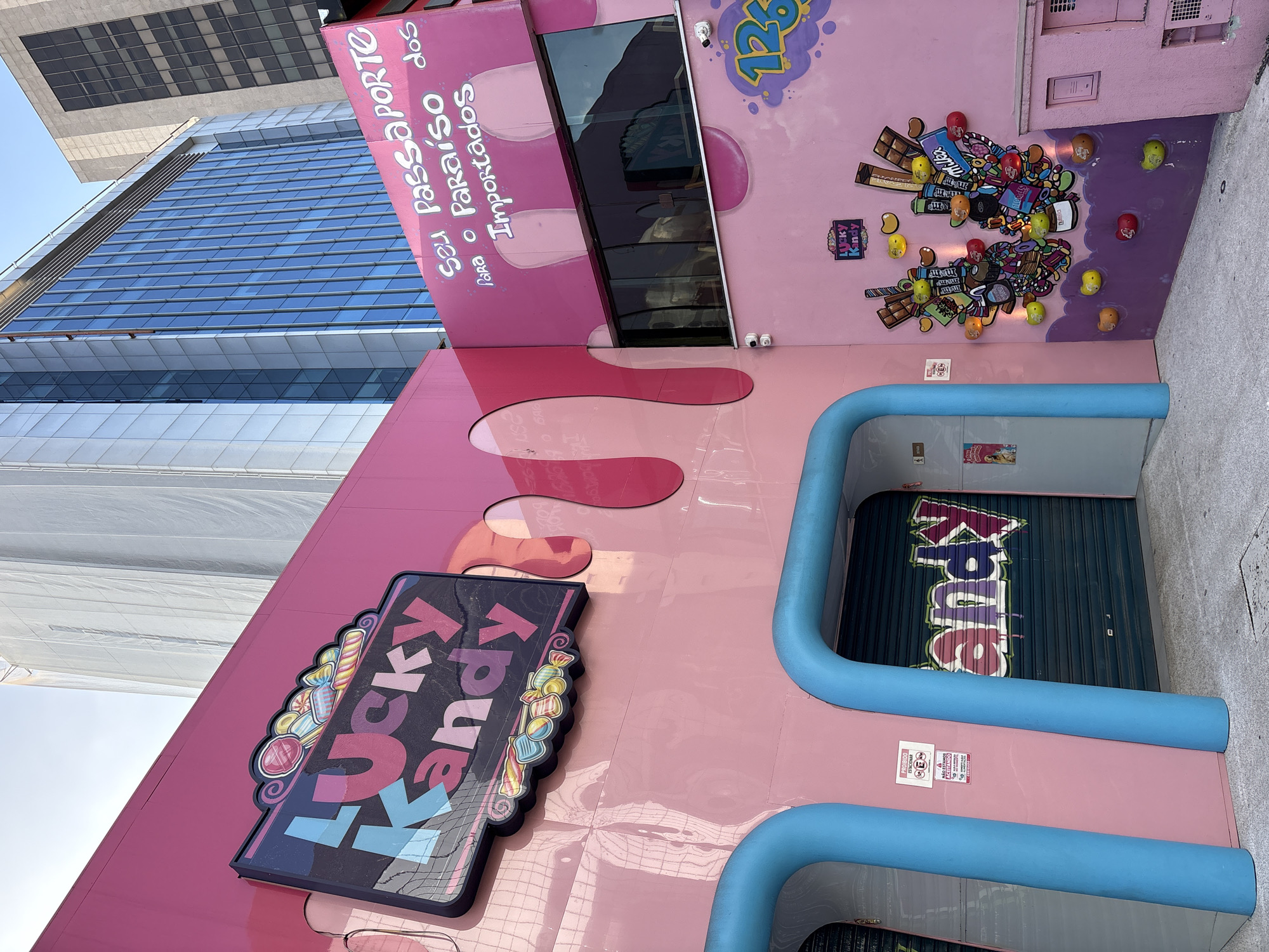 The image shows a vibrant pink building facade in São Paulo, Brazil, a city known for its diverse street art and colorful architecture. The building is decorated with a playful and whimsical theme, predominantly featuring candy and bright colors. A prominent sign on the left reads "Lucky Kandy" in large, colorful lettering with an image of various candies above it. To the right, a smaller sign, also in pink, displays text in Portuguese: "Seu Passaporte para o Paraíso dos Importados," which translates to "Your Passport to the Paradise of Imported Goods." This suggests the establishment is a store selling imported candies.
The building's entrance features two large, curved blue archways leading to a closed metallic shutter with "Kandy" spray-painted in graffiti-style lettering. On the right side of the building, there is a mural depicting a collection of candy-themed objects, including chocolate bars, candy wrappers, and jelly beans, arranged in a decorative manner. A large number "126" is spray-painted in purple and yellow on the upper right corner of this section. The overall aesthetic is one of fun, sweetness, and a touch of urban art. The background reveals modern city buildings with glass facades, indicating an urban setting within São Paulo. There are no people visible in the image.