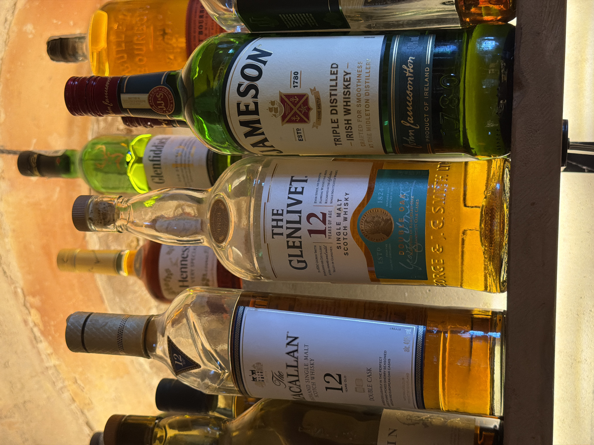 A medium shot shows a collection of alcoholic beverage bottles, primarily whiskey, displayed on a shelf. The lighting is warm and suggests an indoor setting with a textured ceiling above. From left to right, partially visible are a bottle with a dark cap and a gold liquid, followed by a bottle with a black cap and amber liquid. Next is a tall, clear bottle with a blue label that reads "The Macallan 12 Years Old Single Malt Scotch Whisky Double Cask." To its right, a taller, clear bottle with a white and blue label prominently displays "The Glenlivet 12 Years Old Single Malt Scotch Whisky." Further right, a green bottle with a white label features "Jameson Triple Distilled Irish Whiskey Crafted for Smoothness at the Midleton Distillery." A portion of another bottle, possibly also whiskey, is visible to the far right. The overall impression is a curated collection of spirits. The image does not contain any information about the city of Palmanova, Spain.
