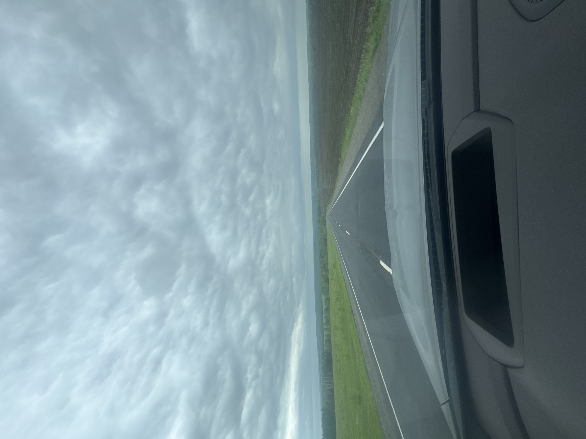 The image shows a view from inside a vehicle, possibly a car or airplane, looking out over a straight, paved road or runway. A dark, cloudy sky is visible to the left of the road/runway. Adjacent to the right of the road/runway is a green grassy field, and further out, a darker cultivated field. In the foreground, parts of the vehicle's interior are visible, including a ventilation opening. The image's orientation is vertical. While the image cannot be directly identified as Новороманово, Russia, without additional information, the visual elements align with a rural environment, which does not necessarily contradict that location.