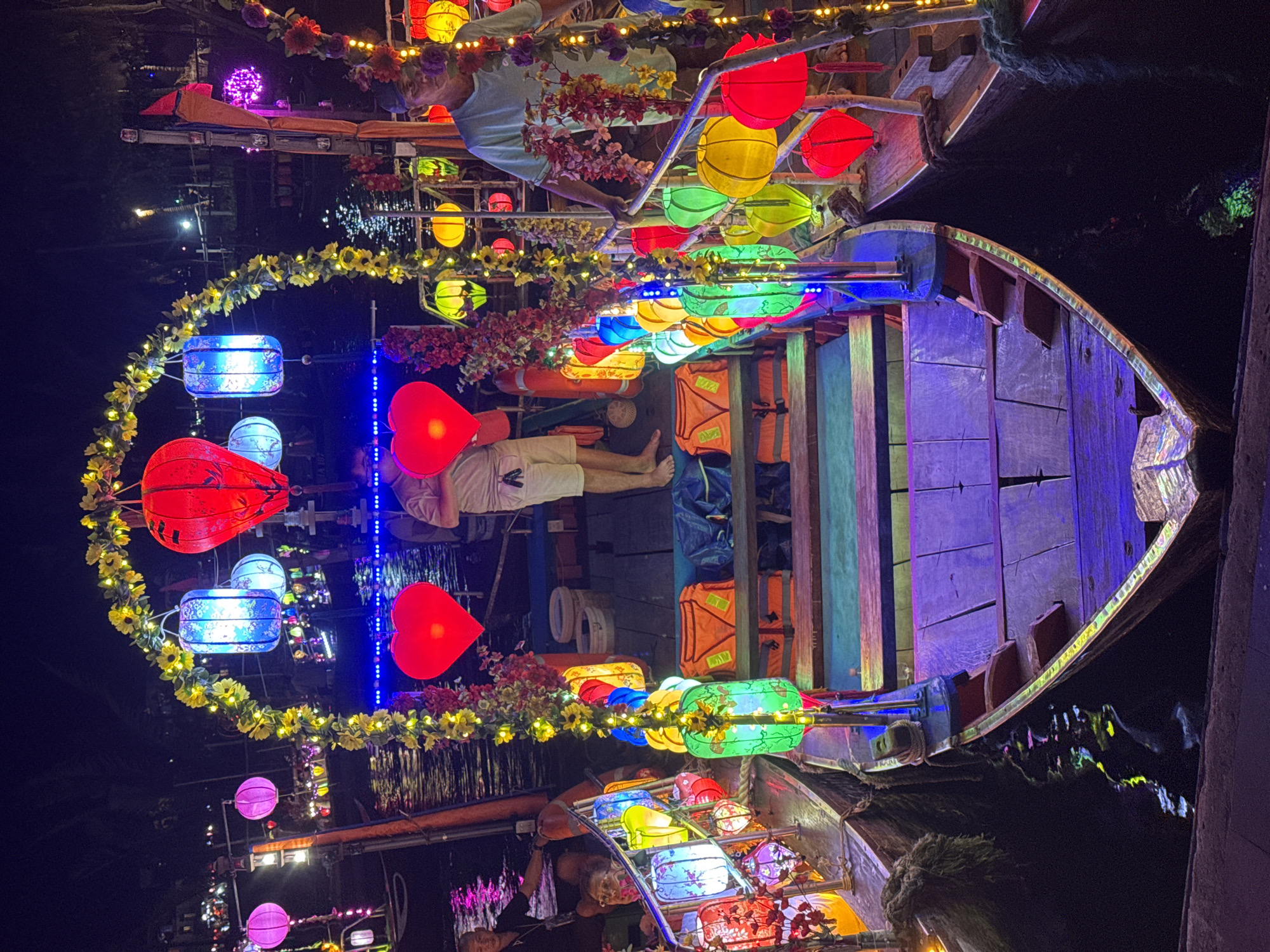 This image depicts a boat at night, decorated with colorful lanterns, likely in Hội An Ward, Vietnam. Two individuals are visible on the boat. One is standing near the front, and another appears to be lying down towards the center. Life vests are also present. Additional boats with similar lantern decorations are visible nearby. The surrounding environment is dark, suggesting the scene is at night.