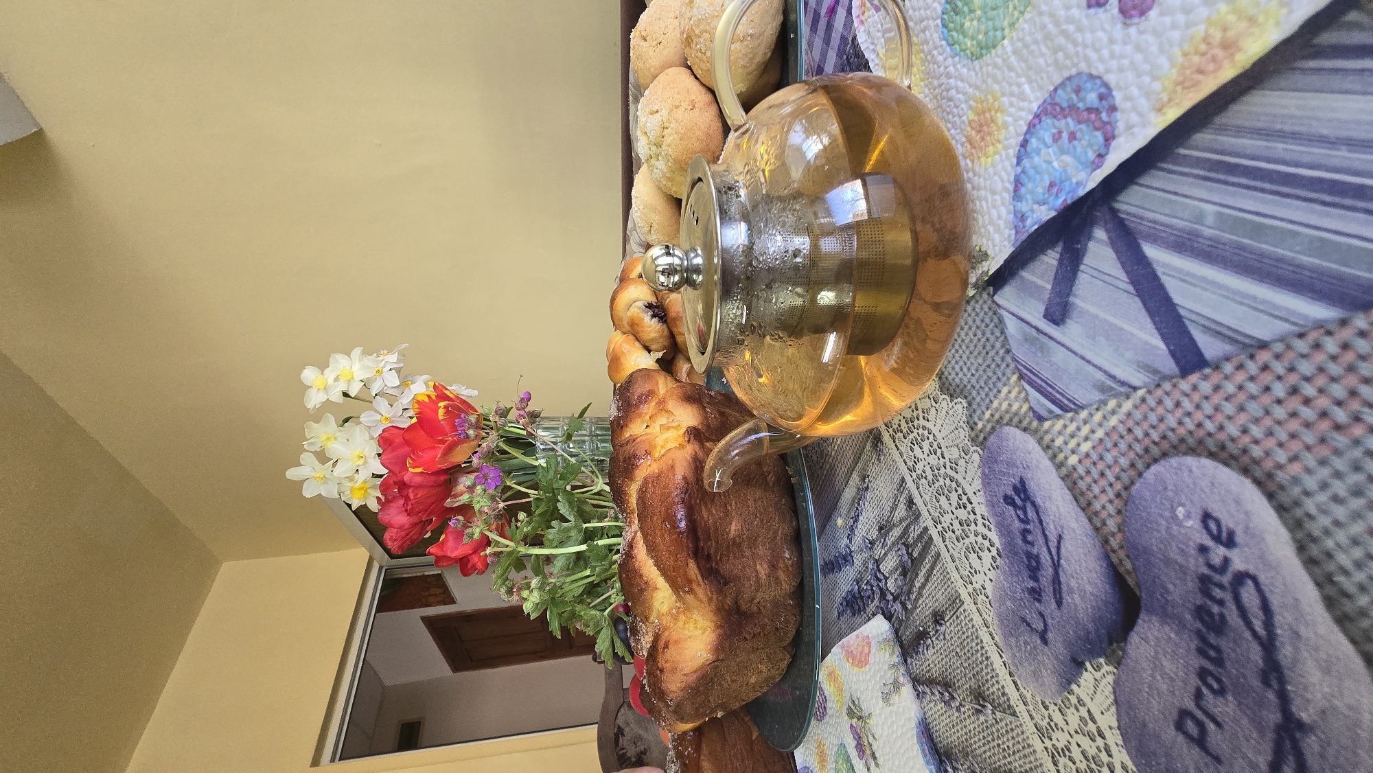 The image shows a still life on a surface covered with different fabrics and textiles. A glass teapot filled with a light amber liquid is positioned on the right, next to a loaf of braided bread on a glass plate and a selection of pastries, including what appears to be blueberry muffins and round bread rolls. A bouquet of white and red flowers is placed to the left. Two lavender-colored items with the words "Lavande" and "Provence" are visible on the surface. The background includes a yellow wall and a glimpse of what might be a window or framed picture. Without further information, it's impossible to confirm the location in Stara Zagora, Bulgaria, or any specifics regarding people, actions, or interactions.