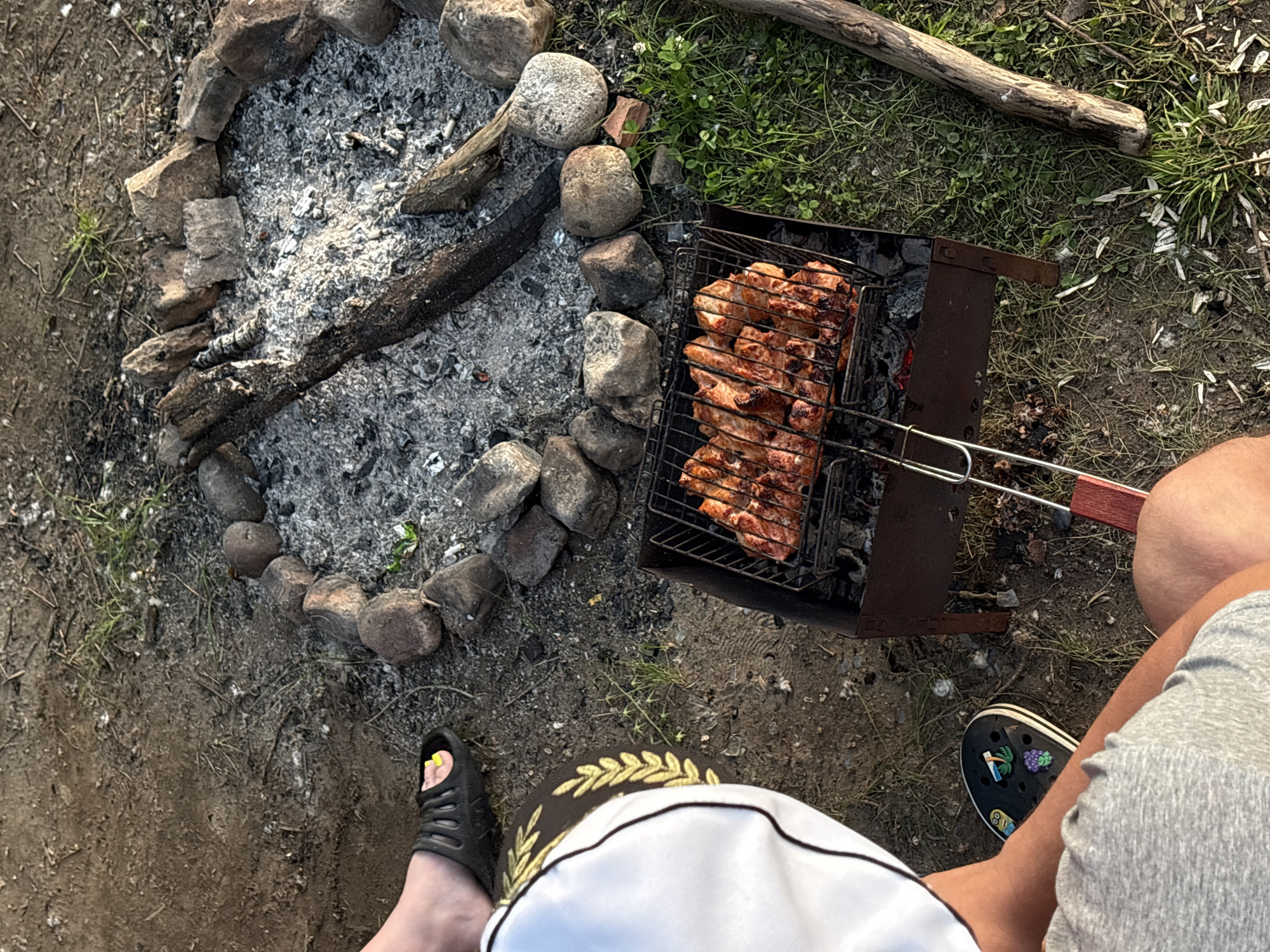 The image depicts an outdoor scene with a barbecue and a campfire. In the foreground, two people are partially visible. One person is wearing grey shorts and a black Croc shoe adorned with various charms, including a minion and a bunch of grapes. The other person is wearing black slide sandals. A white garment with gold trim is also present, possibly clothing.
The main focus of the scene is a barbecue grill loaded with pieces of meat, presumably being cooked over hot coals. Adjacent to the grill is a campfire pit, outlined with stones, containing ashes and burnt wood. The ground is a mix of dirt, grass, and scattered debris, possibly from the campfire. The overall setting suggests a casual outdoor gathering, possibly a picnic or camping trip. The location is described as the city of Perm, Russia, although no specific landmarks are visible in the image to confirm this. The lighting suggests it is daytime, possibly late afternoon or early evening.