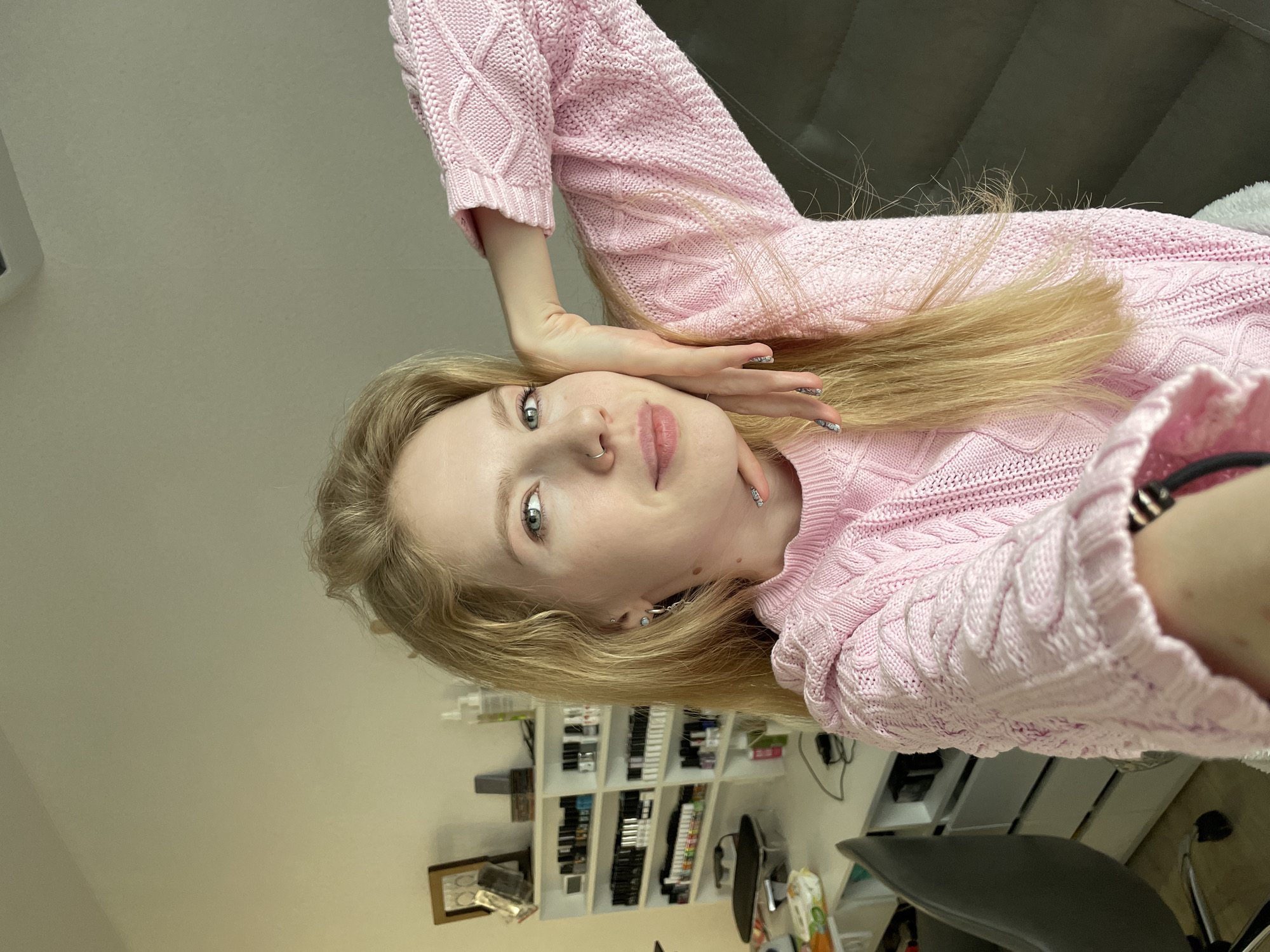 The image displays a young woman with long, straight blonde hair and light-colored eyes, appearing to be lying down and looking directly at the camera with a subtle, soft smile. She is wearing a thick, light pink cable-knit sweater. A small silver nose ring is visible on her right nostril, and she has stud earrings. Her fingernails are adorned with a metallic or glittery silver design. One arm is raised, with her hand gently resting near her forehead and hair.
The background indicates an indoor setting, likely a room that serves as a beauty space or a personal area for beauty enthusiasts. To her right (from the viewer's perspective, assuming the image is rotated upright), a dark grey cushioned surface, possibly a sofa or headboard, is visible, along with a white fluffy item like a blanket or pillow. To her left, a white shelving unit is filled with numerous rows of small bottles, strongly suggesting nail polish or other cosmetic products. Below the shelves, a framed picture can be seen. Further in the background, a dark office chair is partially visible, along with other general indoor items, indicating a lived-in or professional environment. The overall lighting is bright and even, suggesting daytime. The scene conveys a relaxed and casual mood. This image was taken in Izhevsk, Russia.