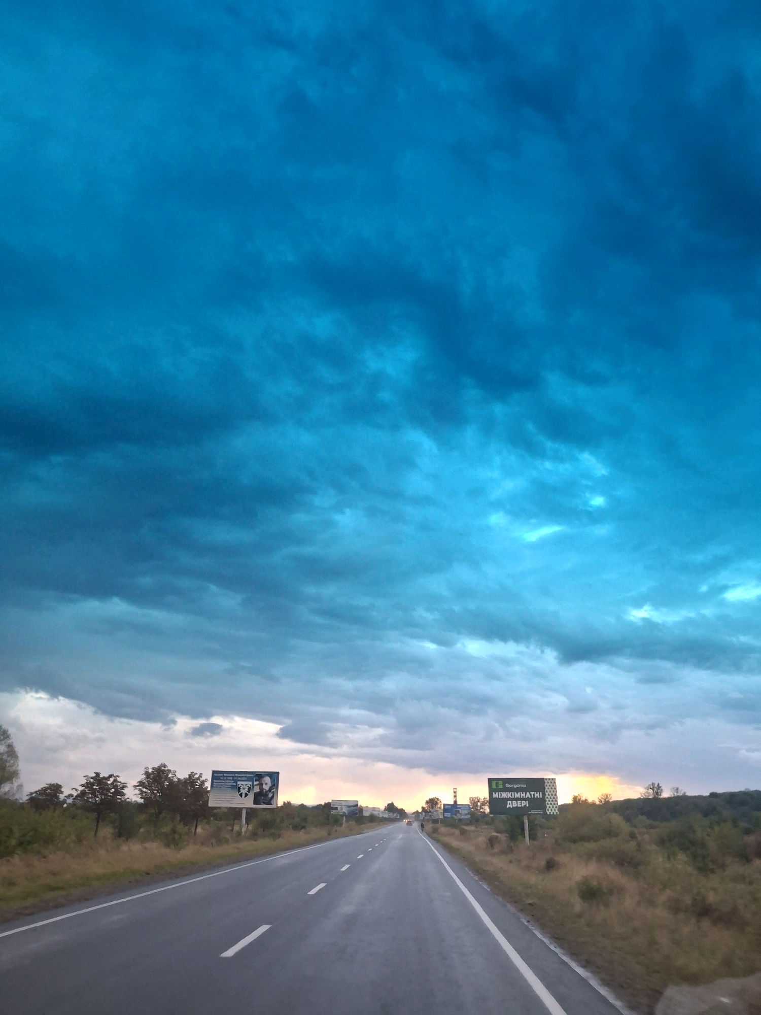 The image shows a road in the vicinity of Piilo, Ukraine, during what appears to be either sunrise or sunset. The sky is filled with dramatic, dark blue clouds, suggesting a potential storm or unusual atmospheric conditions. The road is wet, indicating recent rain. On the right side of the road, there is a billboard with text in Ukrainian that reads "МІЖКІМНАТНІ ДВЕРІ," which translates to "INTERIOR DOORS." Another billboard is visible further down the road on the left, displaying an image of a man and some text. The surrounding landscape is a mix of dry grass, bushes, and scattered trees, with a gentle slope of land on the right. The overall impression is one of a slightly desolate, overcast, and dramatic sky over a rural road.