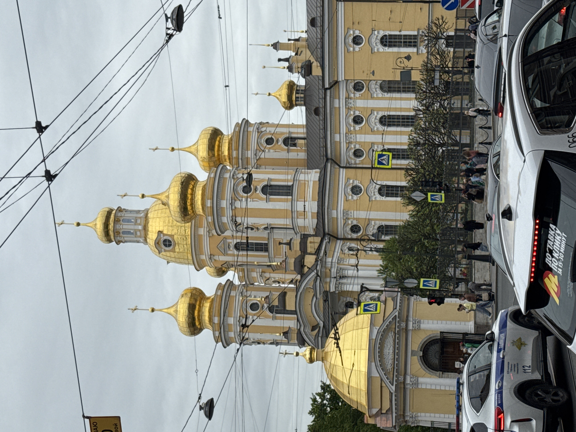 The image captures a scene in Saint Petersburg, Russia, featuring the Smolny Cathedral with its distinctive golden domes as a prominent architectural element. The cathedral's facade is visible behind a busy street scene, where vehicles, including a police car and a taxi, are present. Pedestrians are crossing the street at a crosswalk, marked by yellow and blue signs. Overhead, a network of electrical or tram wires crisscrosses the sky. The overall atmosphere suggests an urban environment with historical architecture juxtaposed against contemporary city life.