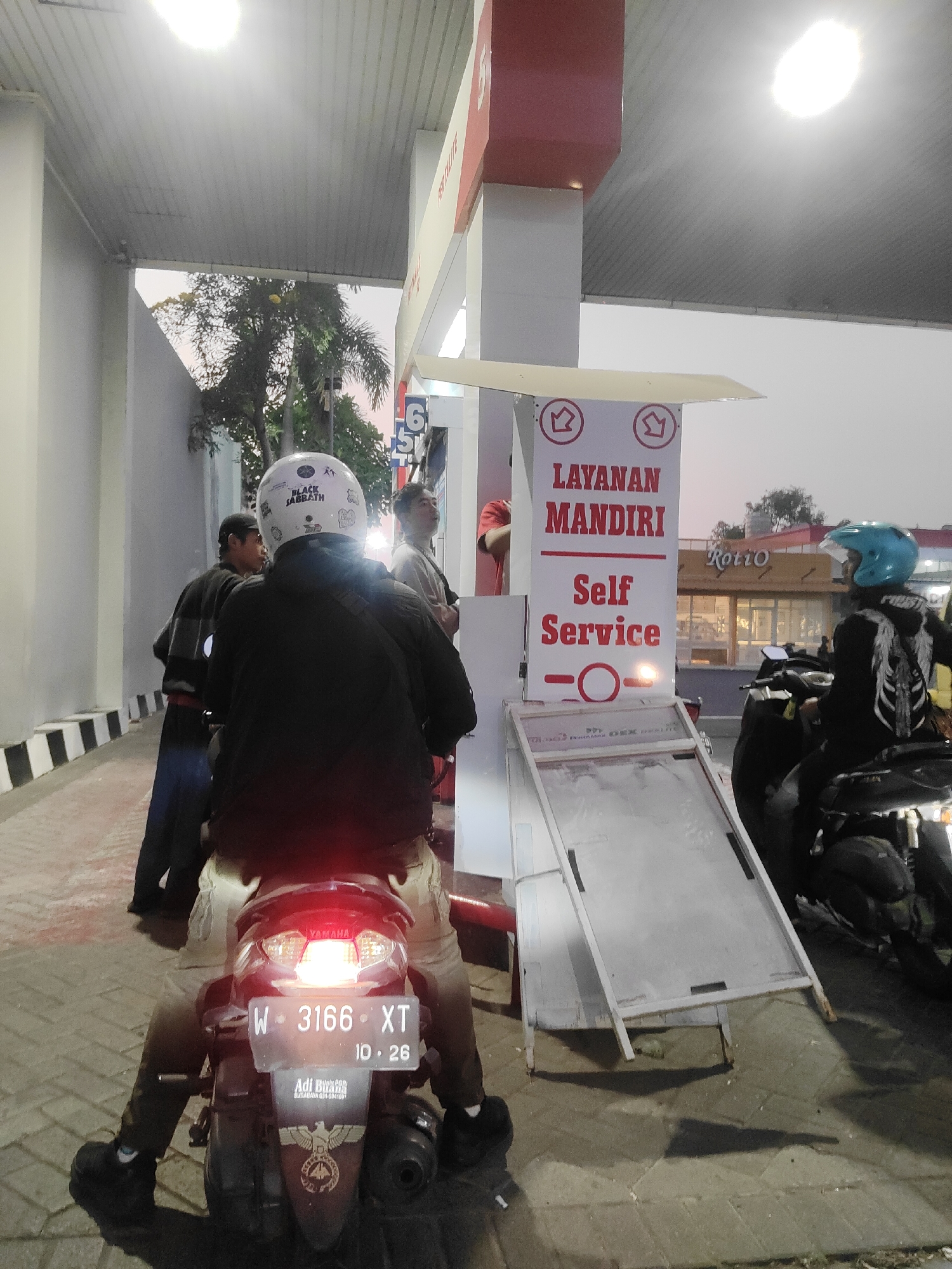 The image shows a street scene at dusk, likely at a gas station. In the foreground, a person wearing a black jacket and a white helmet with "BLACK SABBATH" text is seated on a motorcycle. The motorcycle's license plate reads "W 3166 XT" and "10. 26". The background reveals a white and red gas station structure with signs indicating "LAYANAN MANDIRI Self Service". To the right, another person wearing a blue helmet is on a motorcycle. Several other individuals are visible in the background, with one standing near a kiosk and another person walking. The environment appears to be an urban setting, possibly a commercial area, given the presence of the gas station and a building with "Roti'o" signage. The lighting suggests it is either early morning or late evening. The paved ground has some wet patches and shadows are cast by the structures and people.