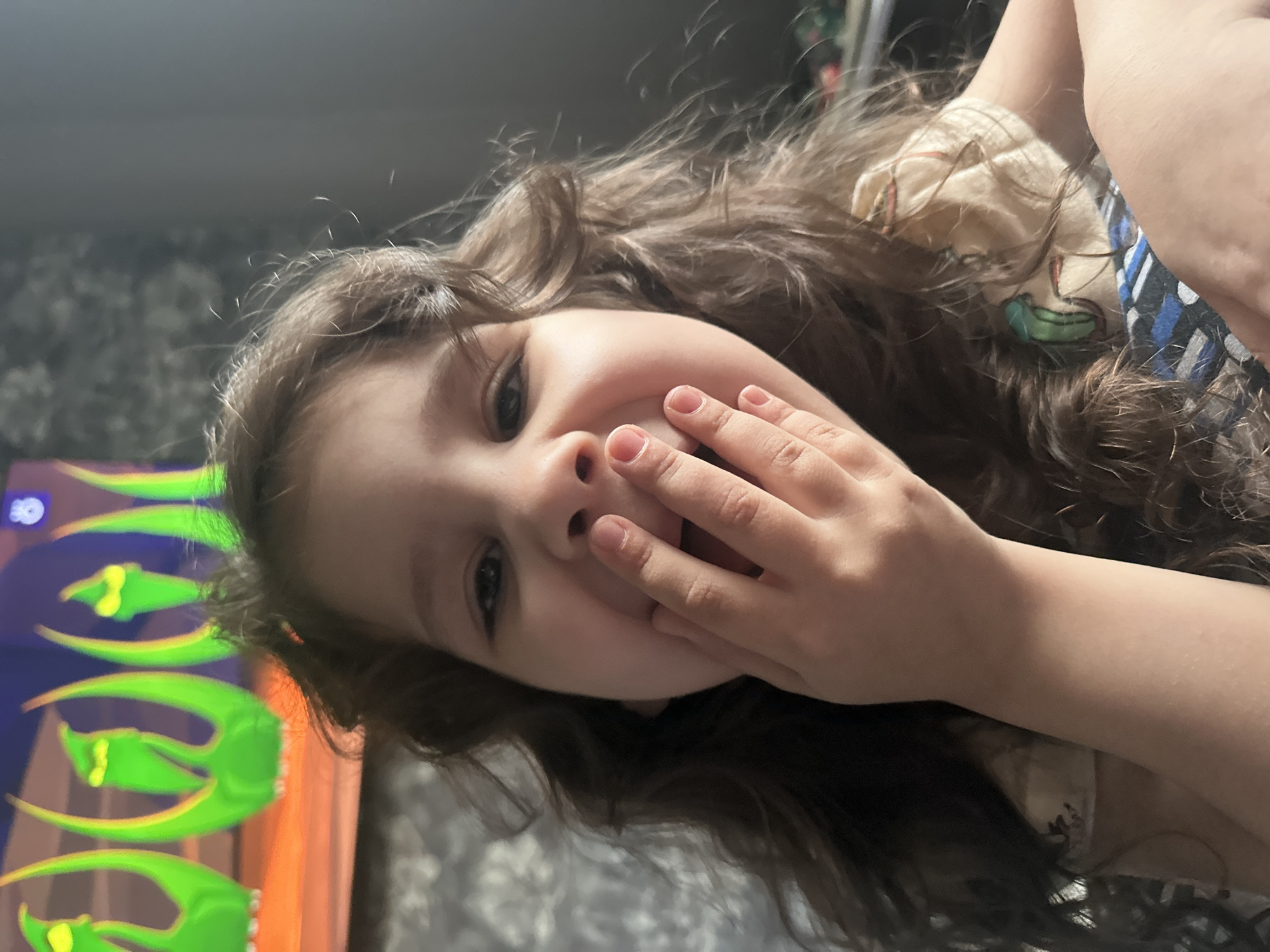 The media file is an indoor, candid portrait of a young child, likely a girl, which is rotated 90 degrees counter-clockwise. The child, with long, wavy brown hair, is looking directly at the camera with a bright, joyful, and possibly shy expression. Her left hand is raised, with her fingers partially covering her mouth, suggesting amusement or bashfulness.
The setting appears to be a home environment. In the background, a television screen displays vibrant, stylized graphics, predominantly bright green and yellow shapes that resemble claws or leaves, against a dark backdrop. In the upper left corner of the screen content, a small white number "30" is visible within a blue circular icon. Behind the child and the television, a dark grey wall with a subtle pattern can be seen. The child is wearing a light-colored garment with faint patterns, and part of a striped top (blue, white, and dark stripes) is visible on her right side. The lighting suggests an indoor setting, and the overall mood of the image is cheerful and spontaneous.