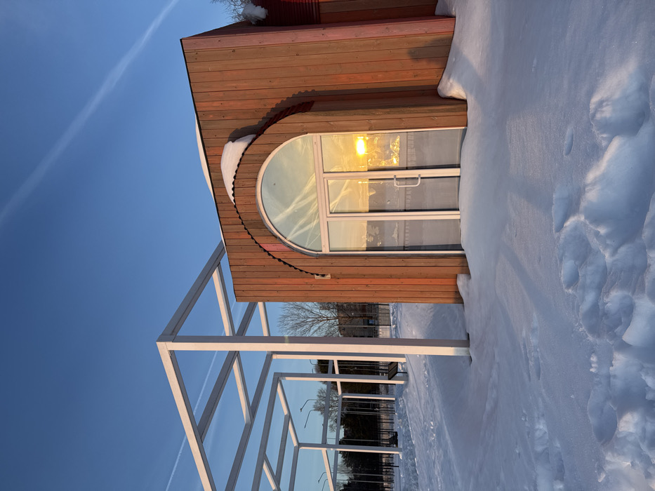 The image captures a winter landscape in Chaikovsky, Russia, during what appears to be late afternoon or early morning, indicated by low-angle sunlight and long shadows. The scene is dominated by deep, undisturbed snow cover blanketing the ground, with distinct footprints visible in the foreground.
In the midground, a standalone building with vertical reddish-brown wooden cladding is present. Its facade features a prominent arched double-door entryway constructed of glass, through which the golden light of the low sun is intensely reflected. A significant accumulation of white snow rests atop the building's roof, particularly above the arched door. To the left of this building, a large, white, modular metal frame structure, resembling a pergola or an architectural skeletal installation, rises from the snow-covered ground, casting sharp, elongated shadows towards the viewer.
The background reveals a row of bare deciduous trees, obscured by the distance and sparse foliage, and hints of other urban structures or fences. The sky above is clear and bright blue, bisected by a distinct white airplane contrail extending diagonally from the upper left to the lower right. No individuals or active subjects are visible within the frame; the scene is static.