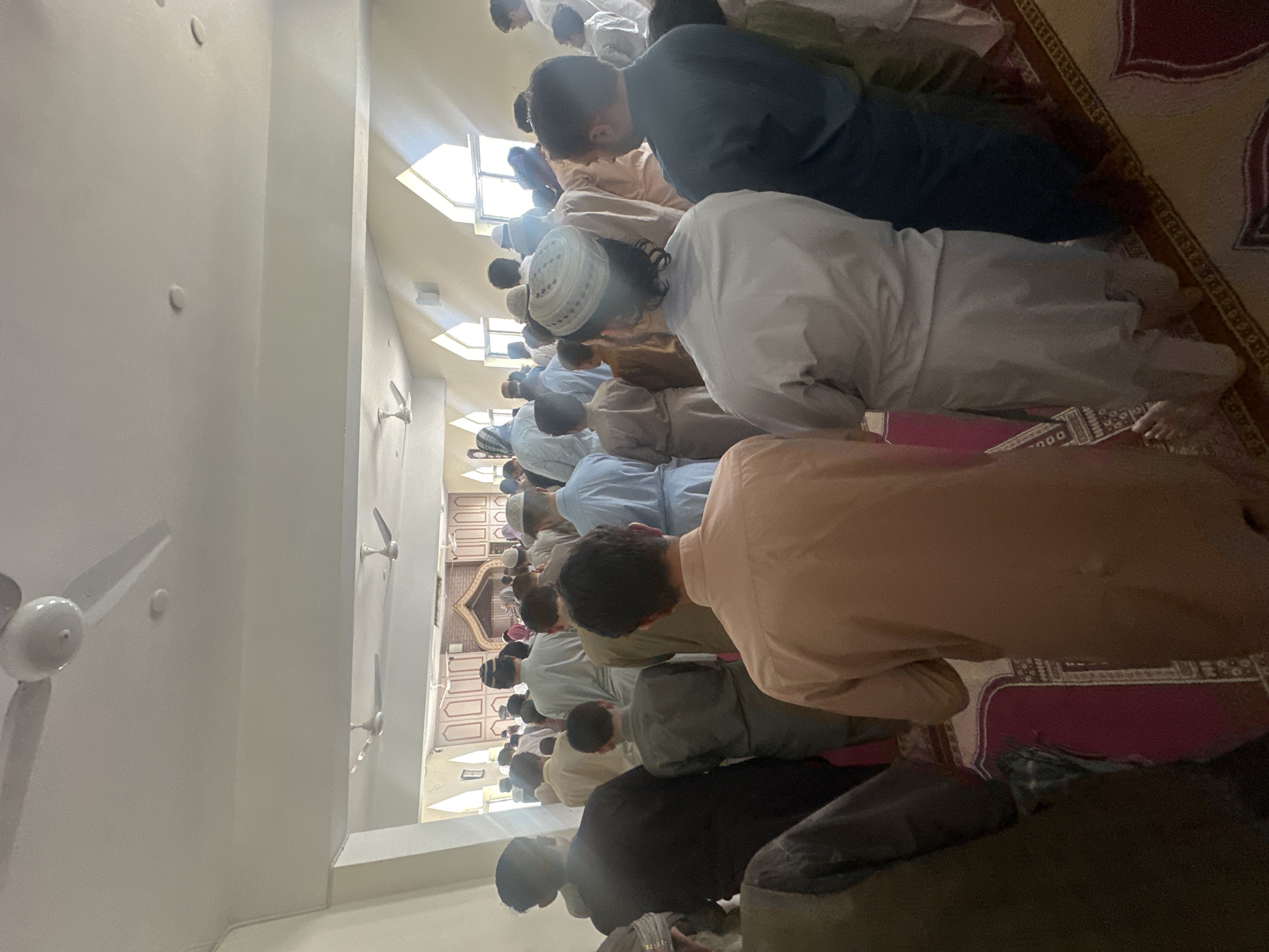 A group of males, varying in age from children to adults, are gathered inside a building, facing the front where a prayer area is visible. They are dressed in traditional Islamic attire, primarily shalwar kameez, in various colors like white, beige, and blue. Some are wearing skullcaps. The scene appears to be a religious gathering, likely a prayer service, taking place in a mosque or prayer hall. The environment is indoors with a ceiling featuring ceiling fans. The walls are painted light cream, and there are arched windows with white frames. A patterned rug covers the floor, and prayer mats are visible in the foreground. The overall lighting suggests it is daytime, with natural light filtering in from the windows. The image captures a moment of collective worship.