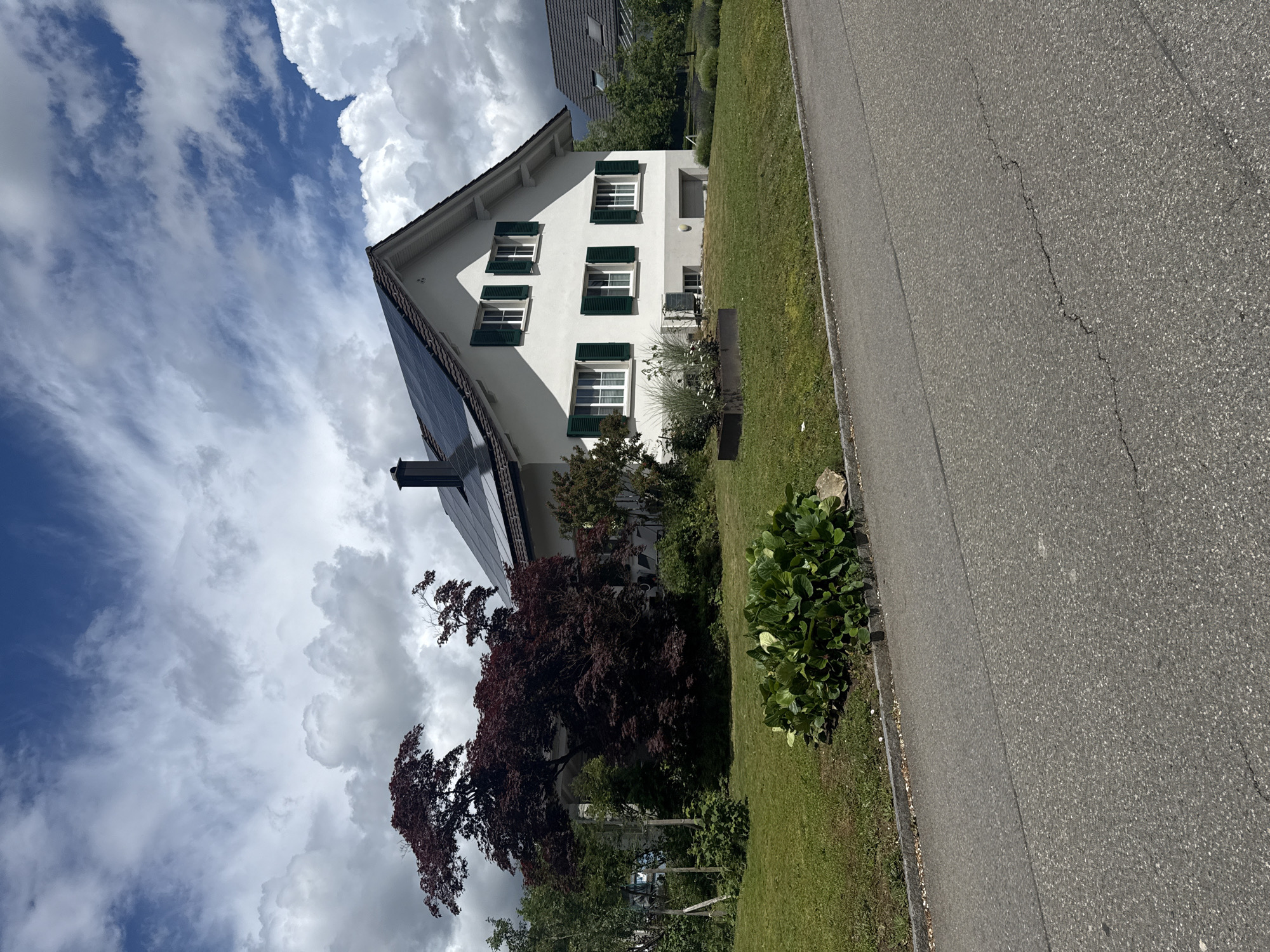 The image depicts a peaceful residential scene in Oberflachs, Switzerland, under a partly cloudy sky. A prominent, two-story white house with a dark-tiled roof stands centrally, featuring multiple windows adorned with dark green shutters. Solar panels are installed on a section of the roof, and a chimney is also visible.
Lush green landscaping surrounds the house, including well-maintained grass verges and various trees and shrubs. A large tree with distinctive dark reddish-purple leaves is visible in the foreground, contributing to the vibrant foliage. To the right of the property, an asphalt road with a visible crack runs alongside, suggesting normal wear. In the background, parts of another house with a dark roof are visible, confirming a residential setting.
The sky is a bright blue, punctuated by large, fluffy white cumulus clouds and some wispy cirrus clouds, indicating clear weather with ample sunshine. The lighting suggests it is mid-day. No people or animals are present, and no specific activity is taking place, contributing to the serene atmosphere. There is no visible text in the image.