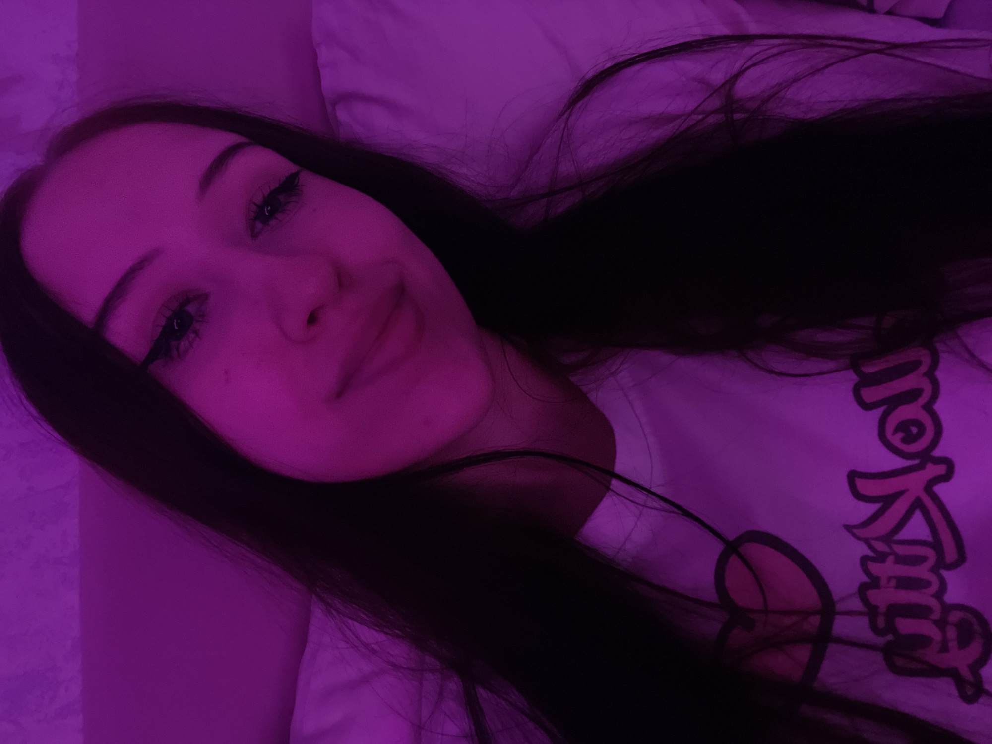 The image is a close-up selfie of a young woman with long dark hair, wearing a white t-shirt with a pink heart and text graphic. She has dramatic winged eyeliner and is looking downwards. The scene is illuminated by a strong pink light, creating a saturated and slightly distorted effect. The background appears to be a bed with a white pillow. There is no discernible indication of the location within Votkinsk, Russia.