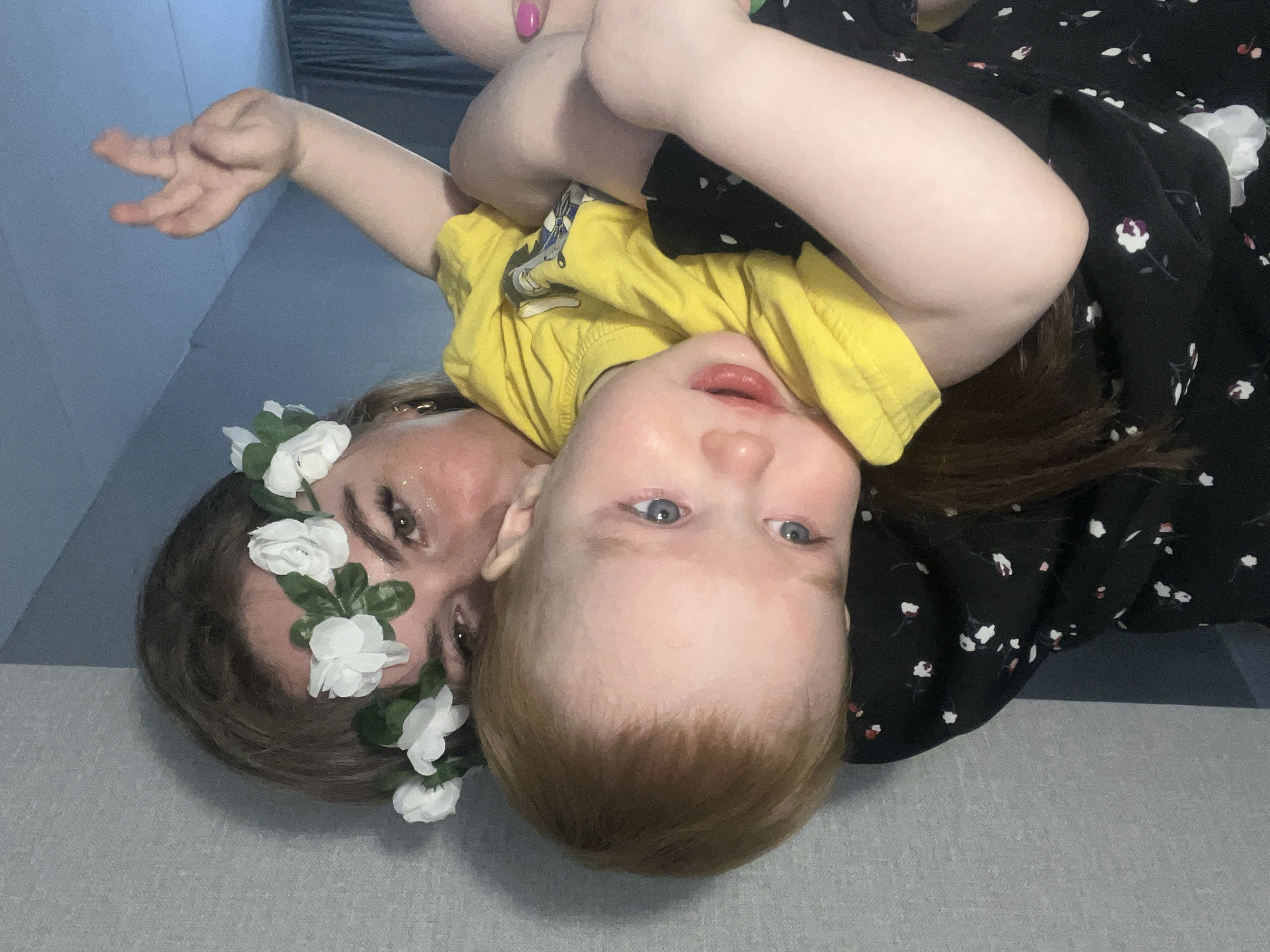 The image shows a woman and a young child in an upside-down orientation. The woman is wearing a flower crown and has glitter on her face. The child is wearing a yellow shirt and is positioned close to the woman. The woman is making a hand gesture, possibly a peace sign. The woman is wearing a black and white floral dress. The background appears to be a gray surface, possibly a floor or bed. The location is not evident based on the provided image. The location in the city of Chaikovsky, Russia cannot be determined from the image.