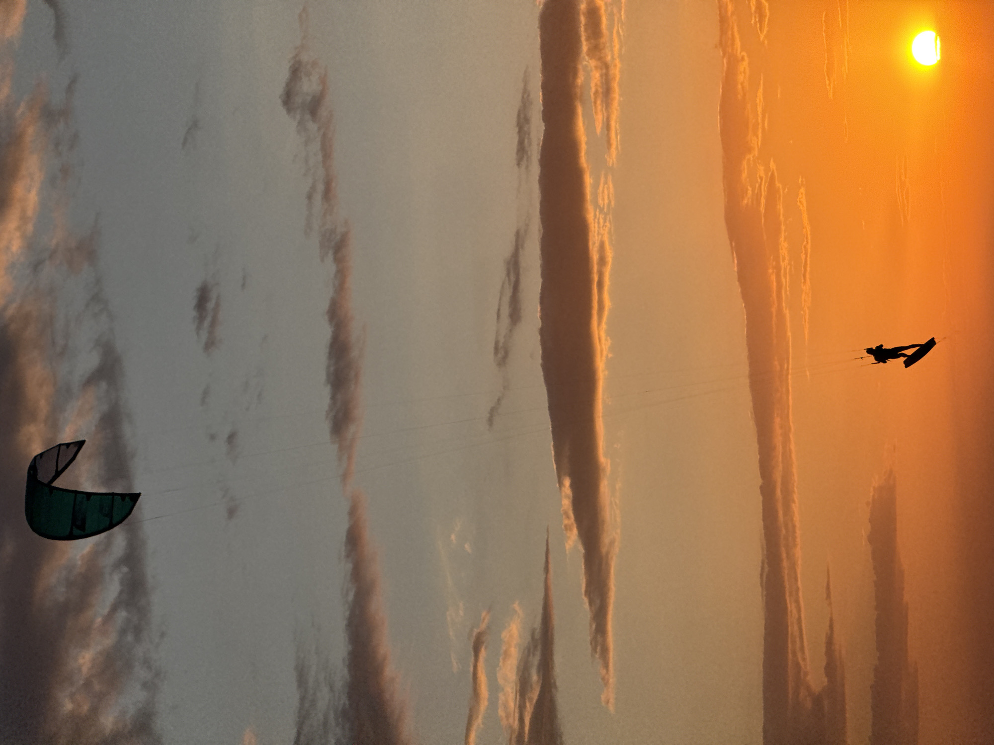 The image is taken in городское поселение Щелкино, Ukraine, during sunset. The sky is filled with clouds and the sun is setting in the upper right corner. There are two kite surfers visible in the sky. One is visible in the lower left corner and the other is near the setting sun.