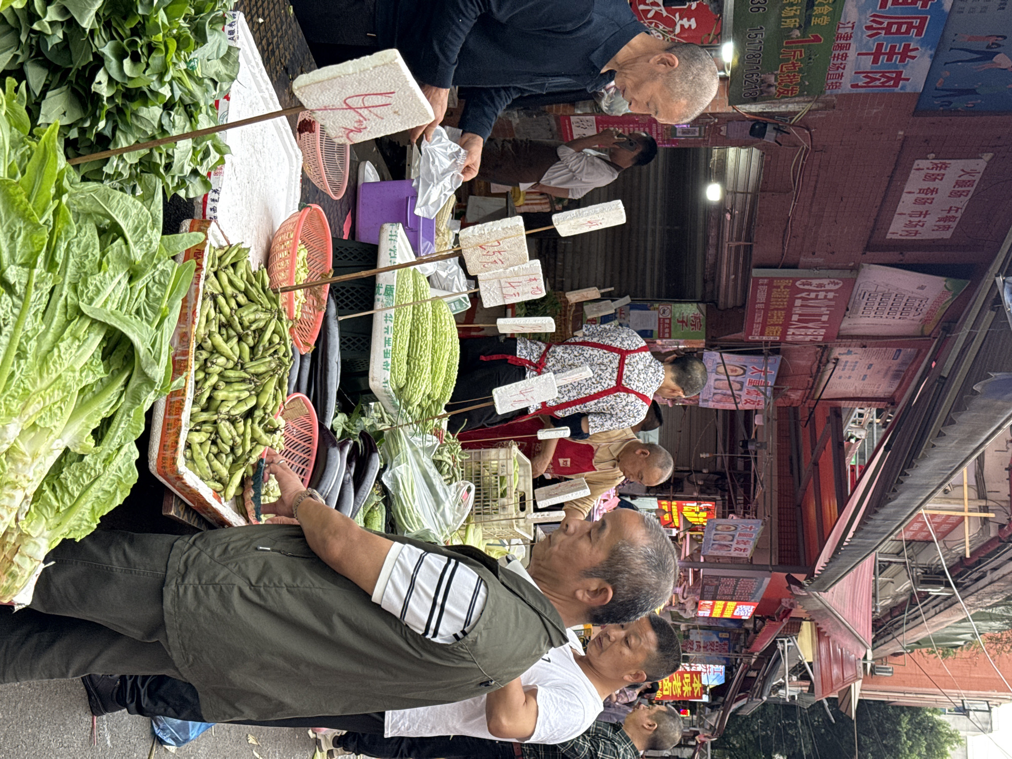The image shows a busy outdoor market in Nan'an District, China. Multiple customers are at a produce stand, examining and purchasing various vegetables including leafy greens, peas, bitter melon and eggplant. Older adults are visible both as customers and vendors. Signs in Chinese characters are posted nearby, advertising food and products. The overall impression is of a bustling, everyday scene of commerce and community.