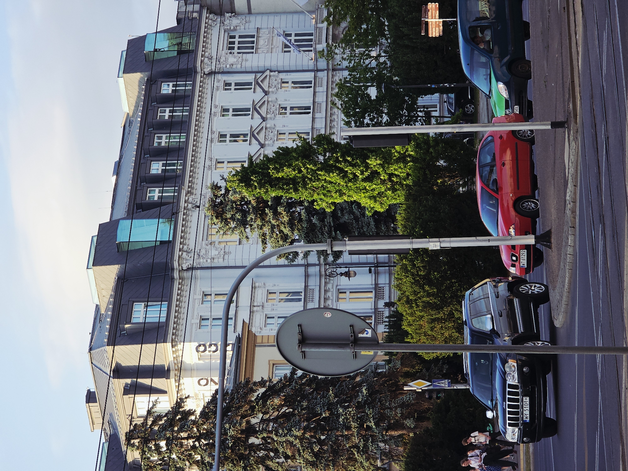 The image depicts a street scene in Warsaw, Poland. The architectural style suggests a location within the city center, potentially near a major thoroughfare or historical district.
Buildings: Multiple multi-story buildings are visible, with a mix of architectural styles. One building appears to be a classic, ornate design, with intricate details and what appears to be a Greek flag hanging on the facade. Another building appears to be a modern structure with a glass exterior or additions.
Vehicles: Several cars of different colors are parked along the side of the road. A Jeep is most prominent and is parked at the center of the photo.
Street Furniture: The scene includes a street sign with a round, gray sign. Poles with traffic lights.
People: A group of pedestrians are partially visible in the lower-left corner.
Environment: Trees and greenery line the street. The sky is visible and appears clear. Tram tracks are embedded into the street.
Overall, the image suggests a street scene within a relatively affluent or central area of Warsaw, where modern and historical architecture coexist alongside typical urban elements like cars and street furniture.