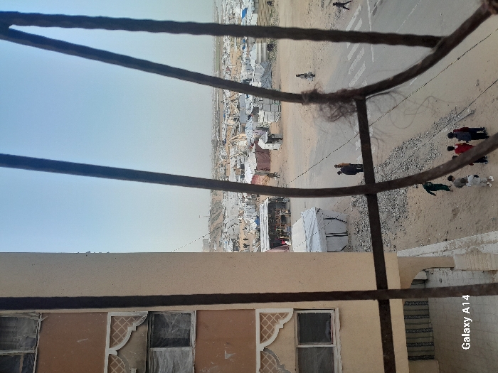 The image presents an elevated view of a densely populated makeshift camp in Khan Yunis, Palestinian Territory, seen through four dark, rusty metal bars, one horizontal bar having fibrous material attached. On the immediate left, a multi-story, light-colored building with decorative lattice patterns between floors features windows covered with clear plastic sheeting. Directly below, a sandy ground leads to a dirt road marked with a white crosswalk. On the road, a group of approximately five children stands on the left, while two adults converse near the center, and two individuals walk further right. Adjacent to the road, a small, open-fronted stall displaying items is visible, alongside several white tent structures. The expansive background is dominated by a vast settlement of numerous white tents and improvised shelters, stretching towards a flat, hazy horizon under a pale blue sky. Distant structures, possibly communication towers, are faintly visible on the horizon.