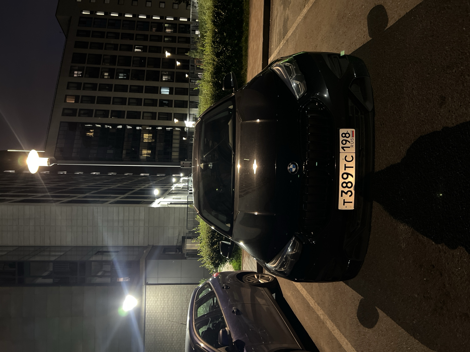 A black BMW is parked in an outdoor lot at night. The car is facing forward, and its license plate reads "T389TC 198 RUS". To the left of the BMW, a portion of a dark blue hatchback is visible. In the background, a multi-story building with numerous illuminated windows stands behind a neatly trimmed hedge. Streetlights illuminate the scene, casting shadows on the asphalt. The location is Saint Petersburg, Russia.