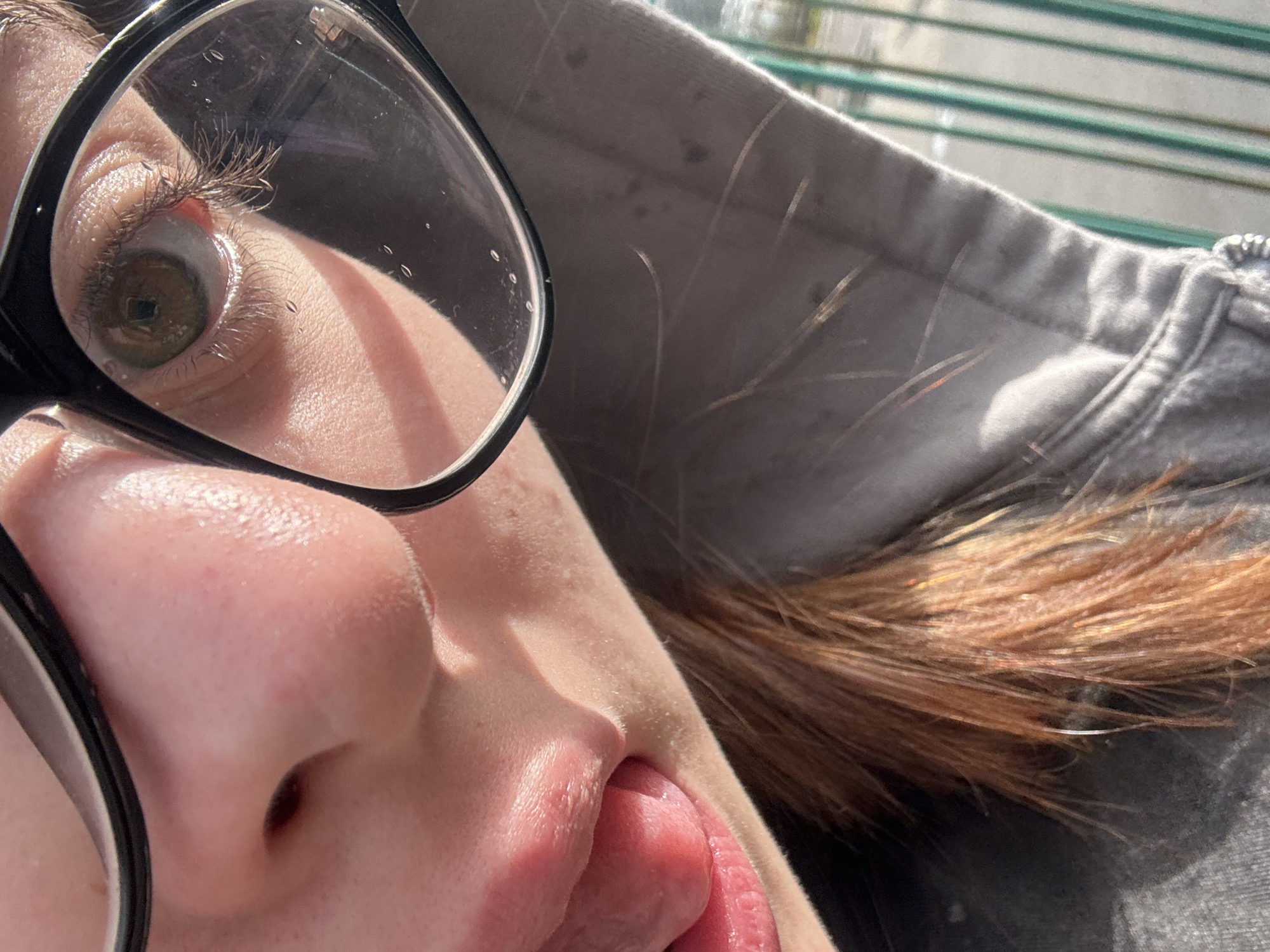 A close-up, distorted perspective image shows a person with their tongue protruding slightly from their mouth. The person is wearing black-rimmed glasses, and their left eye is heavily magnified by the lens. The eye has a hazel or green iris. Skin is pale with visible pores and texture. The person is wearing a grey hooded sweatshirt. Strands of red-brown hair are visible near the hood. The environment appears to be outdoors, with green bars visible in the upper background, likely a railing or fence. The image does not provide enough details to establish the location in the city of Troyan, Bulgaria.
