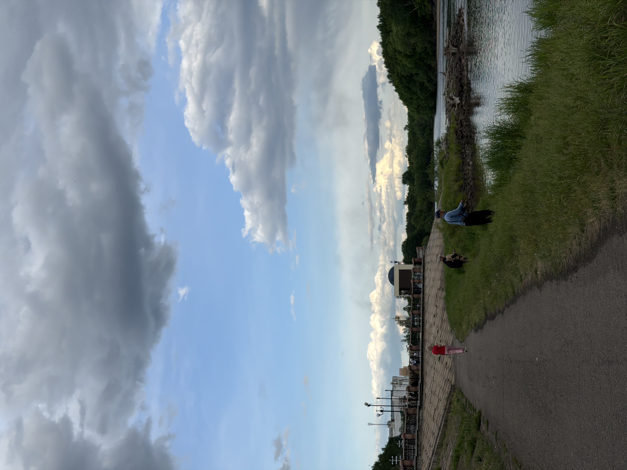 This image captures a wide outdoor scene in Birobidzhan, Russia, depicting a river or lakefront area under a dynamic sky. The perspective is slightly elevated, looking down towards a paved pathway and a body of water.
The sky occupies a large portion of the frame, characterized by a dramatic mix of heavy, dark grey clouds and clear blue patches, suggesting changing weather conditions or a time near sunrise/sunset. The clouds vary in density, with some forming distinct layers and shapes.
In the foreground, a dark paved path stretches diagonally from the lower right to the mid-left. A person wearing a red top and light pants walks along this path, moving away from the viewer. To the left of the path, a decorative railing with streetlights runs parallel, bordering what appears to be a more developed area with parts of buildings visible, including one with a dark, domed roof.
Further to the right, a grassy bank slopes down to a body of water, which appears calm and reflects the sky. Near the water's edge, a person in a blue top and dark trousers is actively fishing with a rod. Close by, another individual is seated on the grass, observing. The bank along the water's edge has some rocky elements and driftwood. On the opposite side of the water, a dense line of green trees forms the distant bank, extending towards the horizon.
The overall scene conveys a sense of peaceful leisure activity in a developed natural setting, likely a park or promenade, during what appears to be late afternoon or early evening given the lighting. No discernible text is visible within the image.