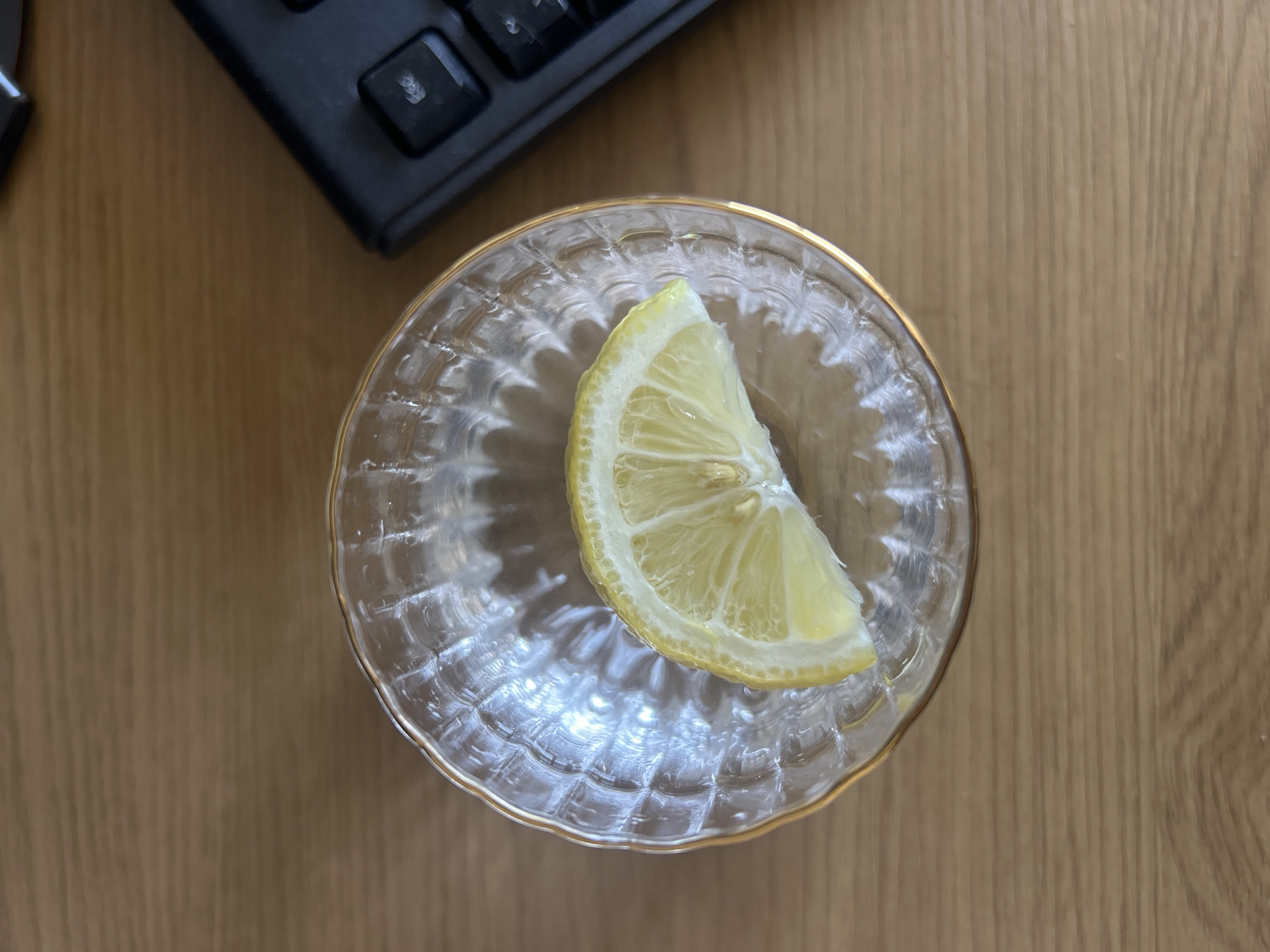 A top-down shot shows a slice of lemon inside a small, clear glass bowl with a gold rim, sitting on a light brown wooden surface. A portion of a black keyboard is visible in the upper left corner of the frame. The lemon wedge is yellow and appears fresh. The bowl has a textured, wavy pattern. The scene is indoors.