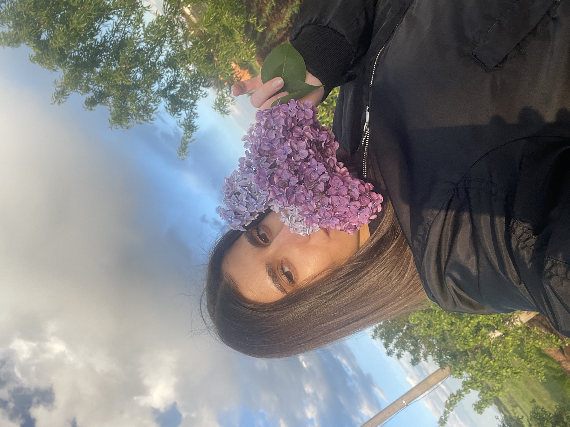 A portrait photograph captures a woman holding a bunch of purple lilacs close to her face, partially obscuring her nose and mouth. She is wearing a black bomber jacket and her dark hair falls towards the bottom of the frame. The background consists of a blue sky with scattered clouds and green foliage, likely trees. The scene is possibly located outdoors, although the specific location within Sofia, Bulgaria is not discernible from the image.
