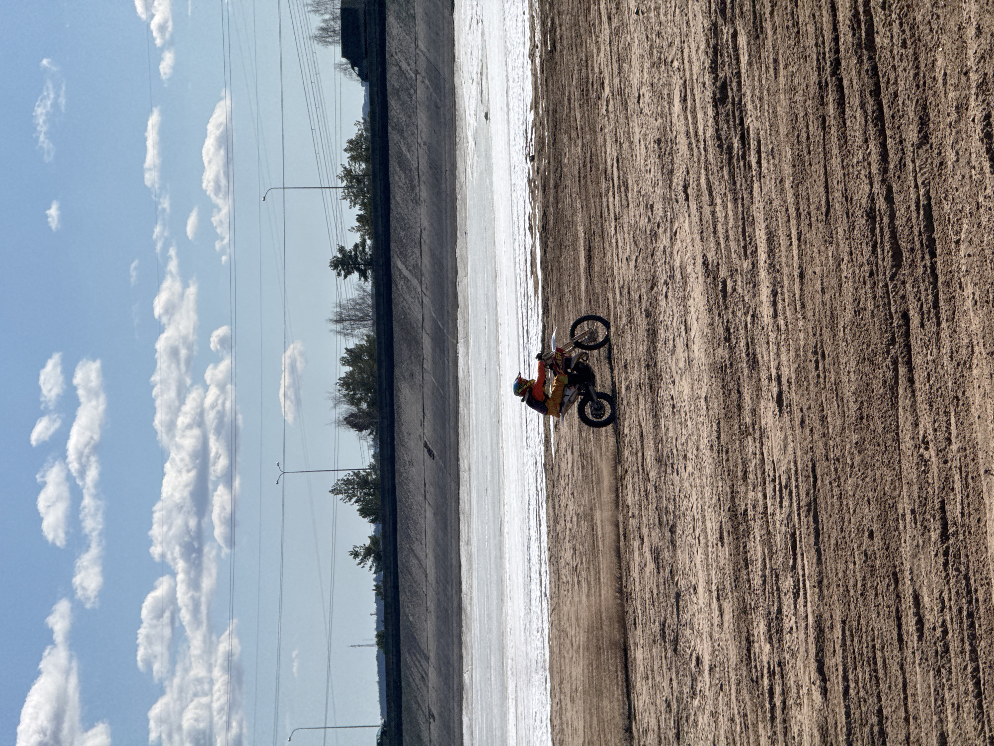 A dirt bike rider maneuvers along a dirt track in Chaikovsky, Russia. The track parallels a concrete channel or canal, with a blue sky and puffy white clouds visible in the background. Power lines and sparse vegetation line the opposite side of the channel. The ground is dry and uneven.