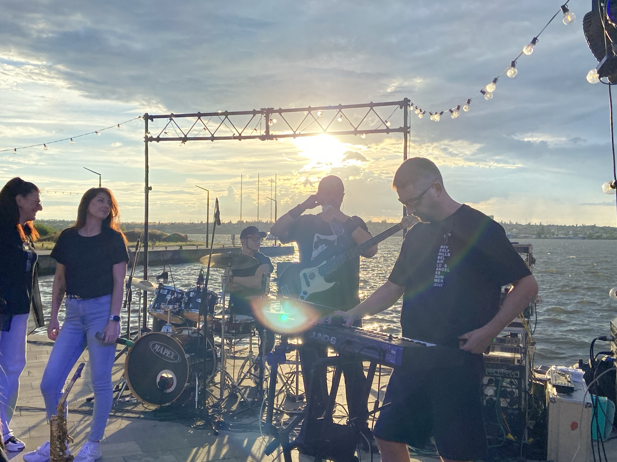 A band performs on a pier at sunset. The sky is filled with clouds and the sun is shining brightly, creating a warm glow. There are several people on stage, including a guitarist, a bassist, a drummer, and a keyboardist. There are also two women standing on the left side of the stage, observing the performance. In the background, there is a body of water and a city skyline. The scene is illuminated by string lights that hang above the stage.