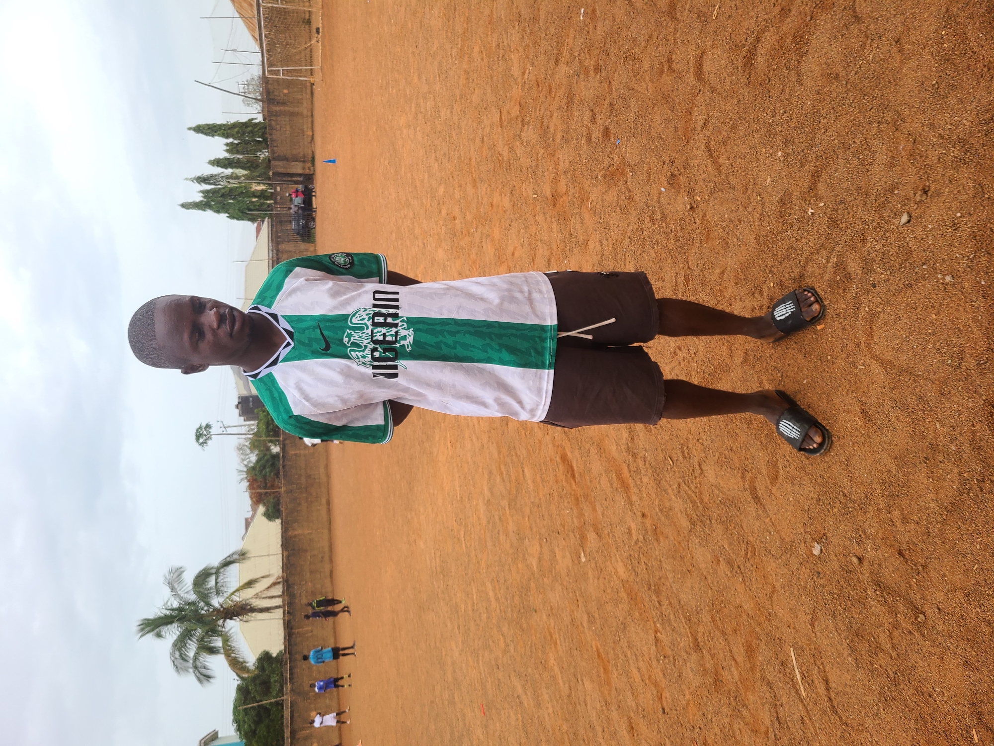 A person is standing on a reddish-brown sandy surface. The person is wearing a white and green shirt with "NIGERIA" printed on it, dark shorts, and black sandals. In the background, there is a wall, trees, and a cloudy sky. In the distance, there are also other people. The location is in the city of Unknown, Unknown.