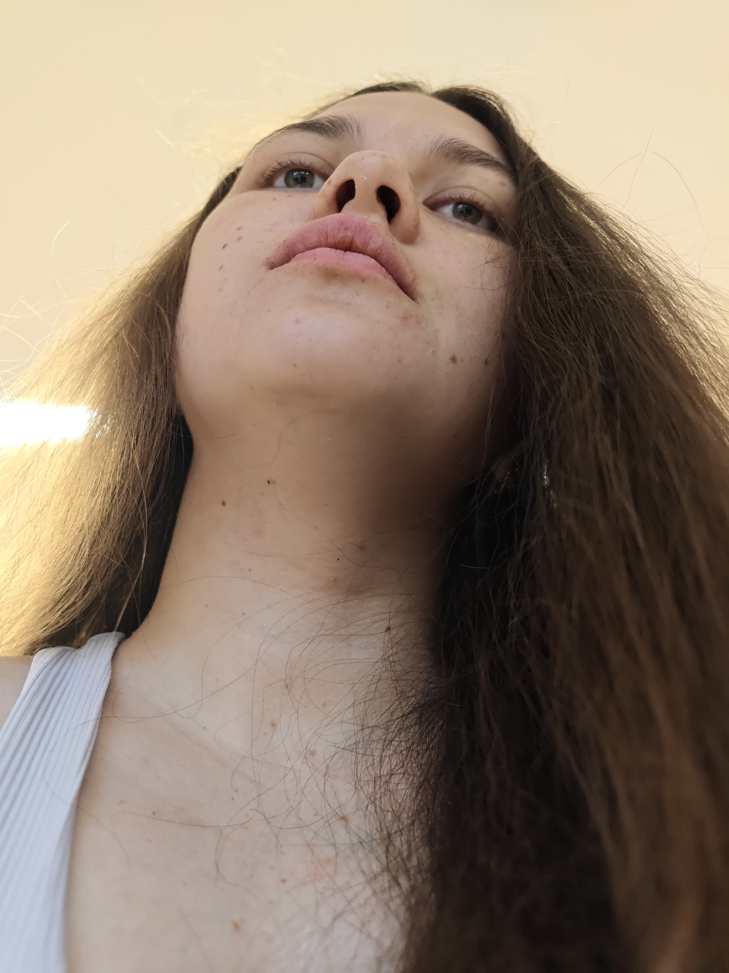 A low-angle, close-up shot shows a young woman with light skin, brown hair, and freckles, looking upwards. Her hair frames her face and falls over her shoulders. She has light-colored eyes and pink lips. She wears a white ribbed tank top. The background is a blurred, light-colored ceiling. The location is Chaikovsky, Russia.