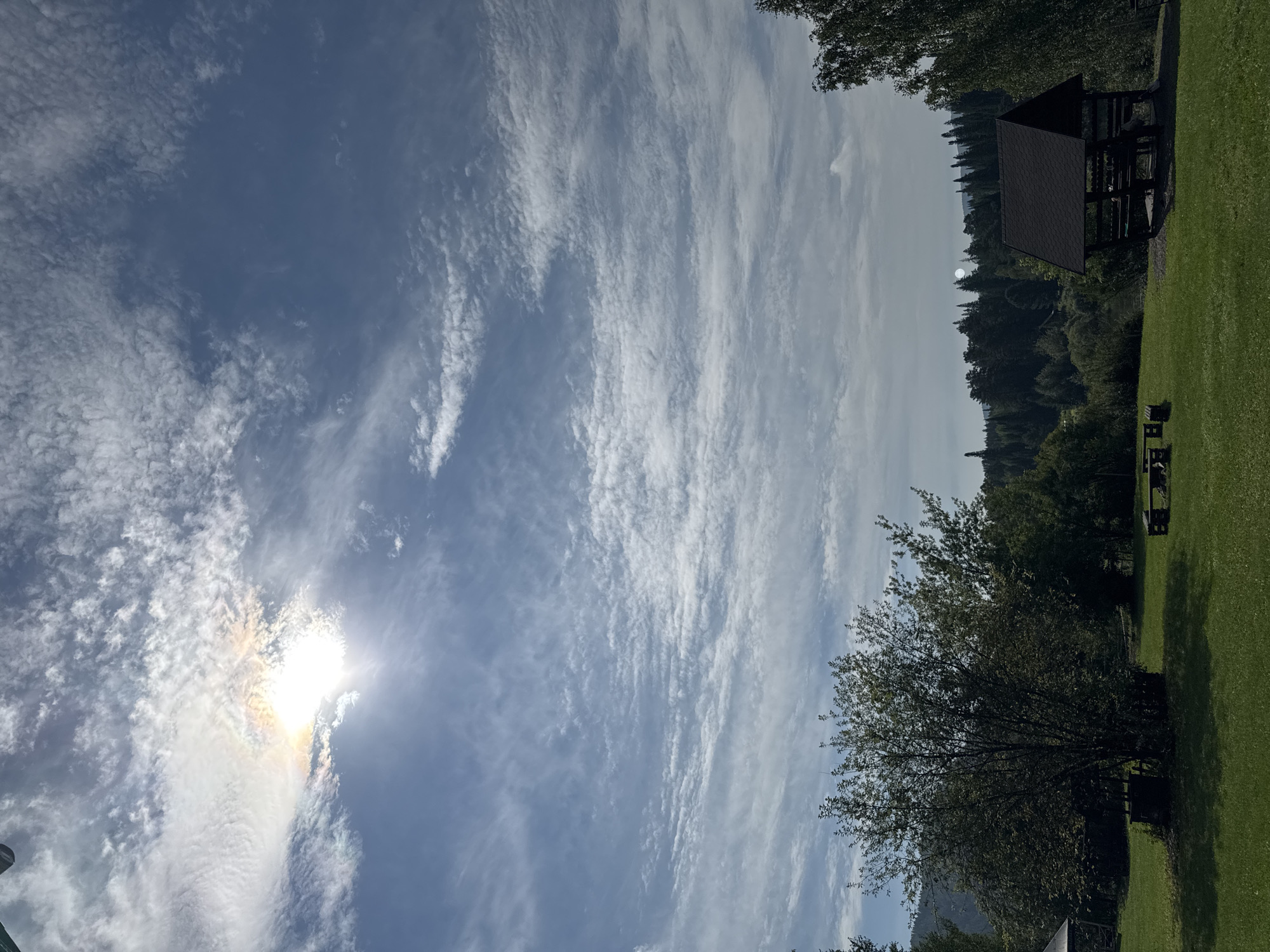 The image depicts an outdoor scene with a wide expanse of sky and a grassy clearing in the foreground. The sky is a vibrant blue, with scattered white clouds, some of which are wispy and others more textured, creating a sense of depth and movement. The sun is visible in the upper left quadrant, partially obscured by a cloud, casting a bright, iridescent halo.
The foreground consists of a well-maintained green lawn. In the middle ground, several elements are present: to the left, a large, leafy tree stands prominently. Near its base, there is a dark, square structure, possibly a well or a decorative element. Further to the right, a dark-roofed gazebo or shelter with open sides and dark posts is situated. In front of the gazebo, there are wooden benches and a picnic table. Behind these elements, a dense forest of dark green trees stretches across the horizon, with rolling hills and mountains visible in the distance. The overall impression is a serene, natural setting, possibly a park or recreational area. The location is identified as Усть-Мана, Russia.