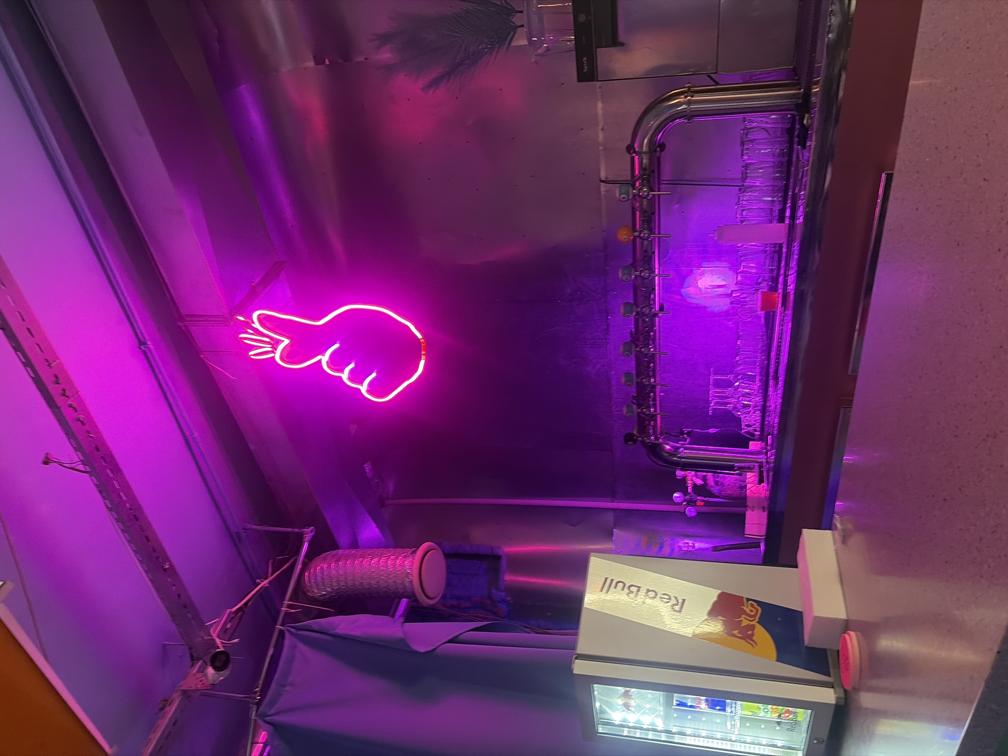 The image depicts a dimly lit bar interior bathed in vibrant pink and purple neon lighting. A prominent neon sign in the shape of a hand making a finger heart gesture hangs from the ceiling. In the foreground, a polished countertop reflects the ambient light. To the left, a small, transparent-doored refrigerator displays cans of Red Bull. Next to it is a tall, rectangular Red Bull branded box. Behind the counter, a row of beer taps is mounted on a metallic structure. Below the taps, numerous empty glasses are neatly arranged on shelves. The overall ambiance is modern and energetic, suggesting a nightlife venue. The provided context indicates the location is Saint Petersburg, Russia.