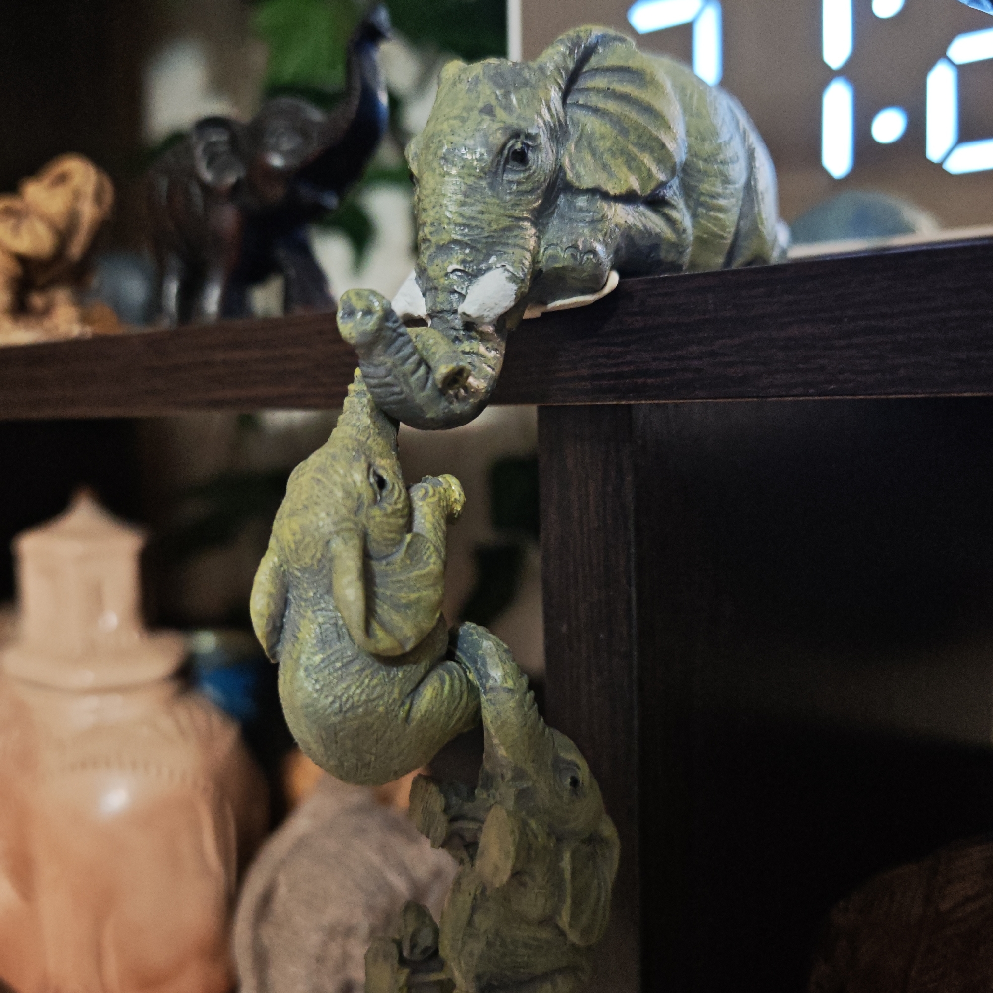 A close-up shot depicts a decorative shelf featuring a set of three elephant figurines. The top figurine rests on the edge of the shelf, while the second appears to be climbing down from the top one. A third elephant figure is at the bottom, supporting the second. The figurines are made of a gray-green material. In the blurred background are other elephant figurines and other objects, including a clock that reads "7:16". The location of Kladovitsy, Russia is not possible to determine from the image.