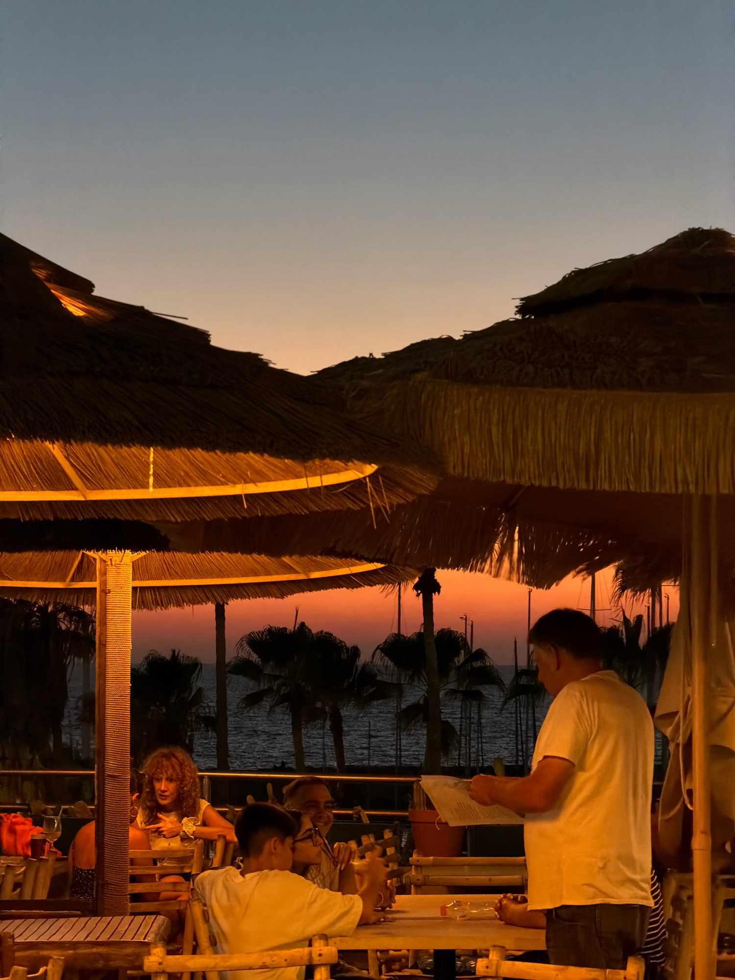 A group of people are dining outdoors at sunset, under thatched umbrellas, at a restaurant located on the Tel Aviv promenade. The sky is a gradient of orange and blue, with the sun having just set below the horizon, casting a warm glow. The sea is visible in the background, with palm trees silhouetted against the colorful sky. A man in a white t-shirt stands at a table, reading a menu. To his left, a woman with curly hair is seated and looking towards the camera. In the foreground, a young person in a white t-shirt is seated at a table, looking down at something on the table. The restaurant appears to have a casual, beachside atmosphere.