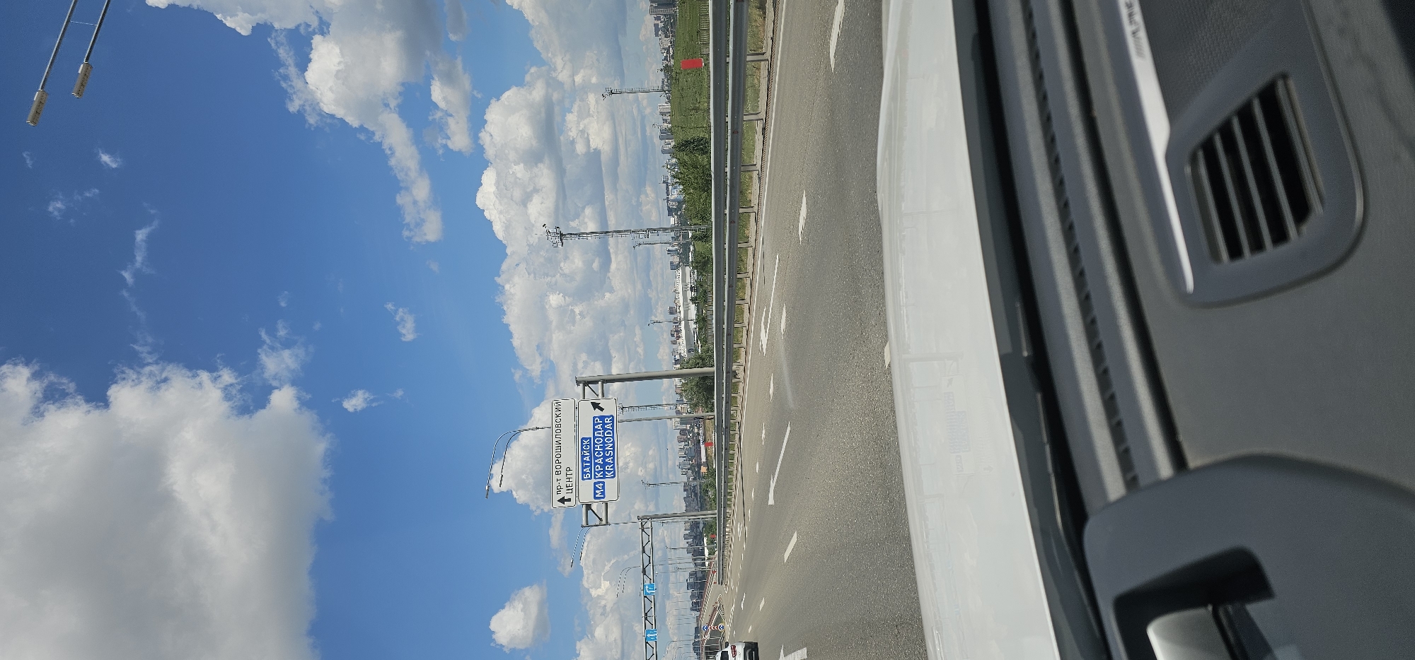 A vertical, eye-level, outdoor shot captures a highway under a partly cloudy blue sky. The view is from inside a white car, looking forward through the windshield. The dashboard of the car is visible in the foreground. In the midground, a multi-lane highway stretches ahead, bordered by a metal guardrail. Road signs are mounted on poles above the highway. The signs are in Russian and indicate directions to "Bataisk," "M4 Krasnodar," and "Voroshilovsky Prospekt," along with "Center." In the background, a city skyline with tall buildings and some industrial structures is visible. There are streetlights along the highway and tall, modern lampposts with multiple lights on poles scattered throughout the scene. The lighting suggests it is daytime, with the sun casting shadows.
