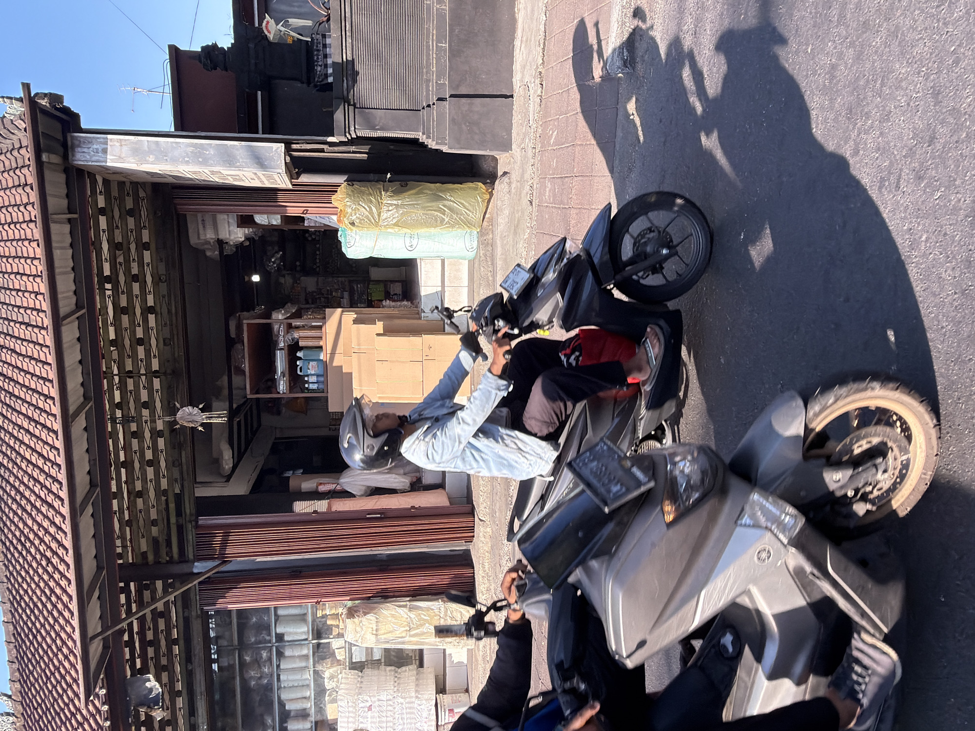 Two people ride motorcycles down a street in North Kuta, Indonesia. The motorcycle in the foreground is silver and its front wheel is visible. The motorcycle in the background is black and is carrying a passenger wearing a light blue jacket and a black helmet. The passenger is looking forward and appears to be riding. The buildings in the background are commercial establishments with open doorways, displaying various goods such as boxes, cylindrical items wrapped in plastic, and bottles of liquid. The sun is shining brightly, casting long shadows on the paved road and sidewalk. The architecture suggests a tropical climate, with terracotta roof tiles and dark stone pillars visible on the right side of the image.