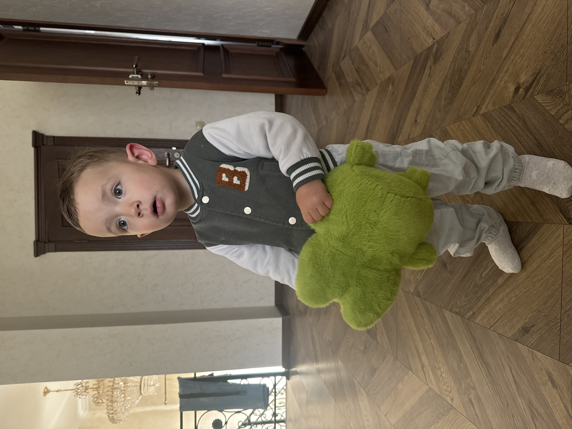 A young boy stands on a wooden herringbone-patterned floor, clutching a green stuffed toy that obscures part of his body. He wears gray pants and a gray and white varsity jacket with a brown patch featuring the letter "B." He looks upward and to the left with a surprised expression. A doorway with dark wood trim and a metal doorknob is visible in the background, along with a wall with a slightly textured surface. In the distance, visible through a doorway or archway, is a chandelier and what appears to be an exterior view of architecture. It is not possible to determine if the location is in the city of Макарово, Russia from the given image.