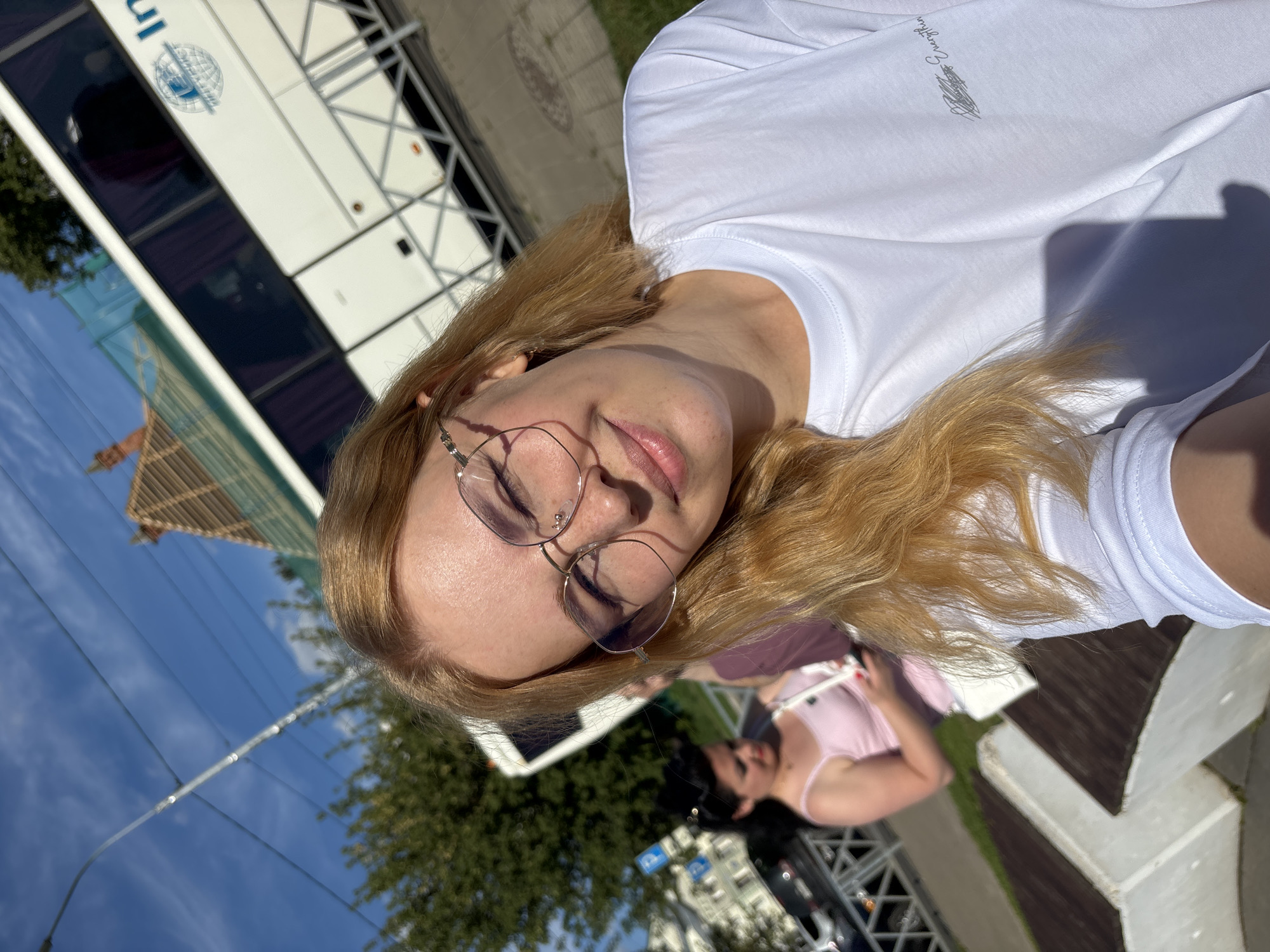 A young woman with blonde hair and glasses is taking a selfie outdoors on a sunny day. She is wearing a white t-shirt. In the background, a white bus with tinted windows is parked. Another woman in a pink top is visible behind the main subject, holding a phone. The location appears to be a street or park area with trees and a blue sky. A street lamp and power lines are also visible. There is no specific information about Rusanovo, Russia visible in the image.