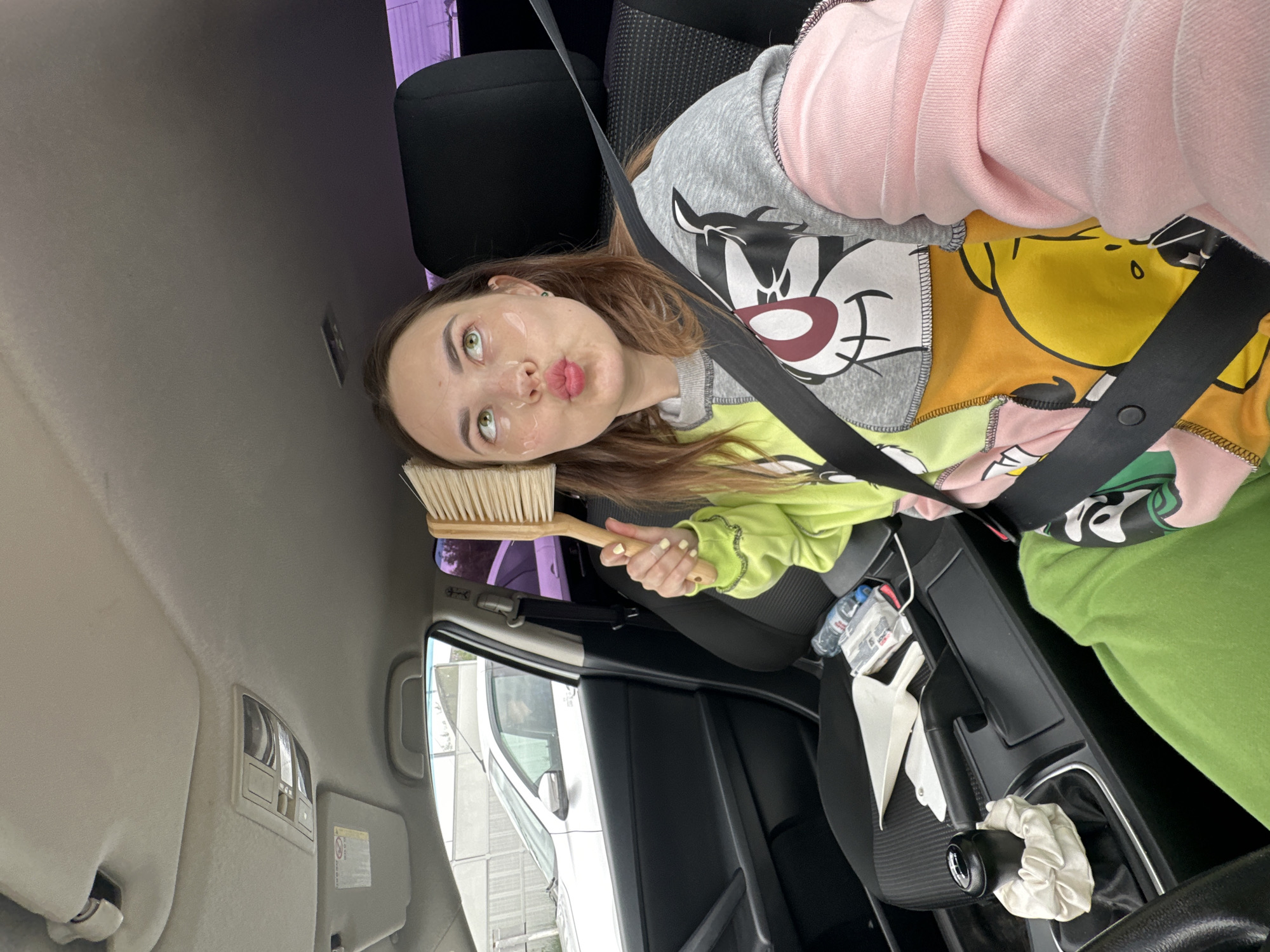The image depicts a young woman sitting in the driver's seat of a car. She is wearing a colorful, patchwork sweatshirt with cartoon characters and a bright green garment on her legs. She has clear eye patches on her cheeks and is holding a wooden-handled brush to her forehead, puckering her lips. The car's interior is visible, including the dashboard, gear shift, and a seatbelt. Outside the car, through the windows, a light-colored car and a fence can be seen. The presence of a car and typical urban elements suggest the location is within a city. However, the provided image does not contain any specific visual cues that would allow for the precise identification of the city as Perm, Russia, or any other specific urban location.