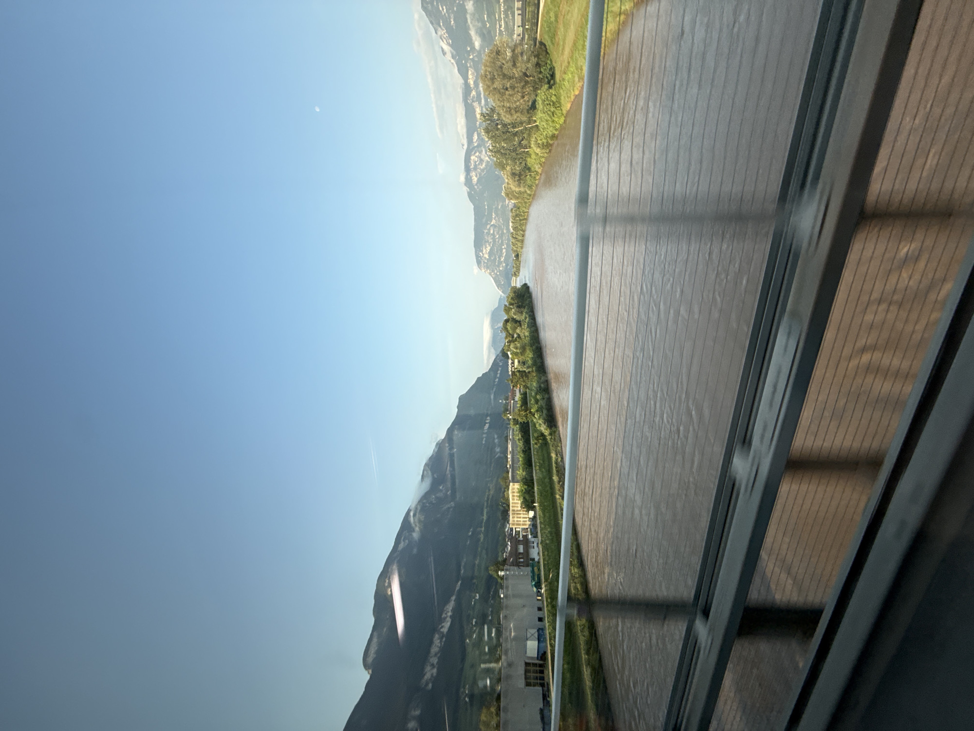 The image shows a wide, muddy river flowing through a valley surrounded by mountains. On the left bank of the river, there are buildings and greenery, indicating a developed area. The mountains on both sides of the valley are covered in vegetation, with some bare rock faces visible. The sky is clear and blue, with a faint moon visible in the upper right corner. The perspective suggests the photo was taken from a bridge or an elevated position, possibly a moving vehicle, given the slight blur in the foreground railing. The overall scene depicts a natural landscape with some human infrastructure integrated into it, likely in the region of Neumarkt - Egna, Italy, given the geographical context of mountainous terrain and river valleys often found there.