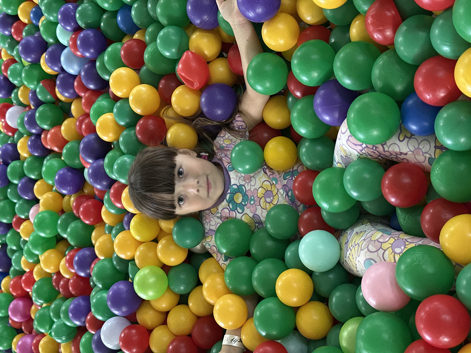 The image depicts a young female child situated within a large ball pit in the city of Chaikovsky, Russia. The child, appearing approximately four to seven years of age, has dark brown hair with bangs and gazes directly forward. She wears a light-colored top adorned with a floral pattern incorporating yellow, purple, and green motifs, with a visible light purple collar. Her torso and head are elevated above the balls, while the remainder of her body is submerged. A white wristband is present on her left wrist. The ball pit is densely populated with numerous spherical plastic balls, primarily green, yellow, red, purple, blue, pink, and white. Several balls exhibit minor superficial indentations or faint manufacturing text. The lighting within the scene is uniform, consistent with an indoor recreational setting.