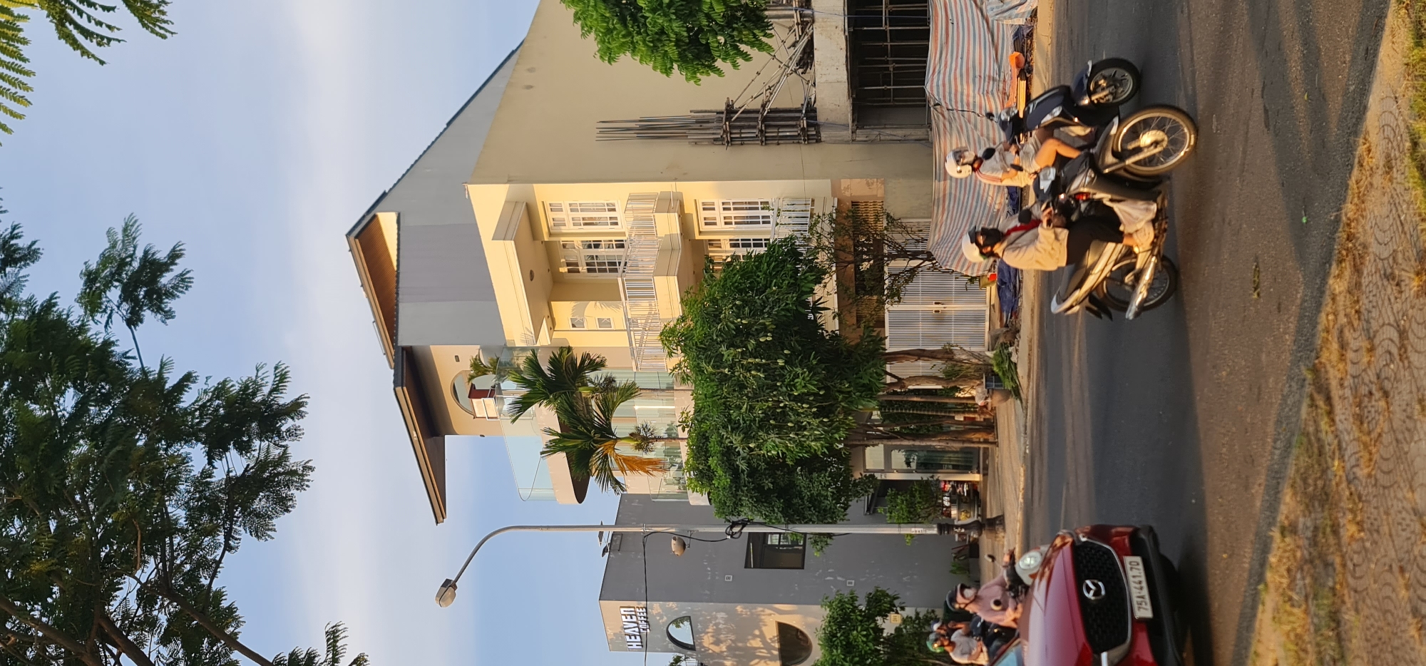 This image captures a vibrant street scene in Đà Nẵng, Vietnam, bathed in the warm light of late afternoon or early morning. The sky is a clear, bright blue, indicating good weather.
The scene features a row of multi-story buildings lining one side of a road. To the left, several modern-looking structures are visible, including one prominent light-yellow building with multiple balconies and white railings. Next to it, a building under construction or renovation shows exposed scaffolding and bare concrete. Further down the street, a grey building with an arched entrance features a sign that clearly reads "HEAVEN." Lush green trees, including tall leafy ones and smaller palm trees, are abundant, providing natural elements and shade along the street and in front of the buildings.
On the road itself, several motorbikes are present. Some are parked, while others have riders wearing helmets, suggesting movement or waiting. One rider is visible on a dark motorbike towards the right, and another on a silver-grey motorbike in the mid-ground. Part of a red car, which appears to be a Mazda CX-5, is visible in the foreground, with its license plate "75A-441.70" clearly readable. A lamppost stands prominently in the mid-ground. The pavement on the right side of the road features a decorative pattern. The overall atmosphere is that of a typical, busy urban street in a tropical setting.