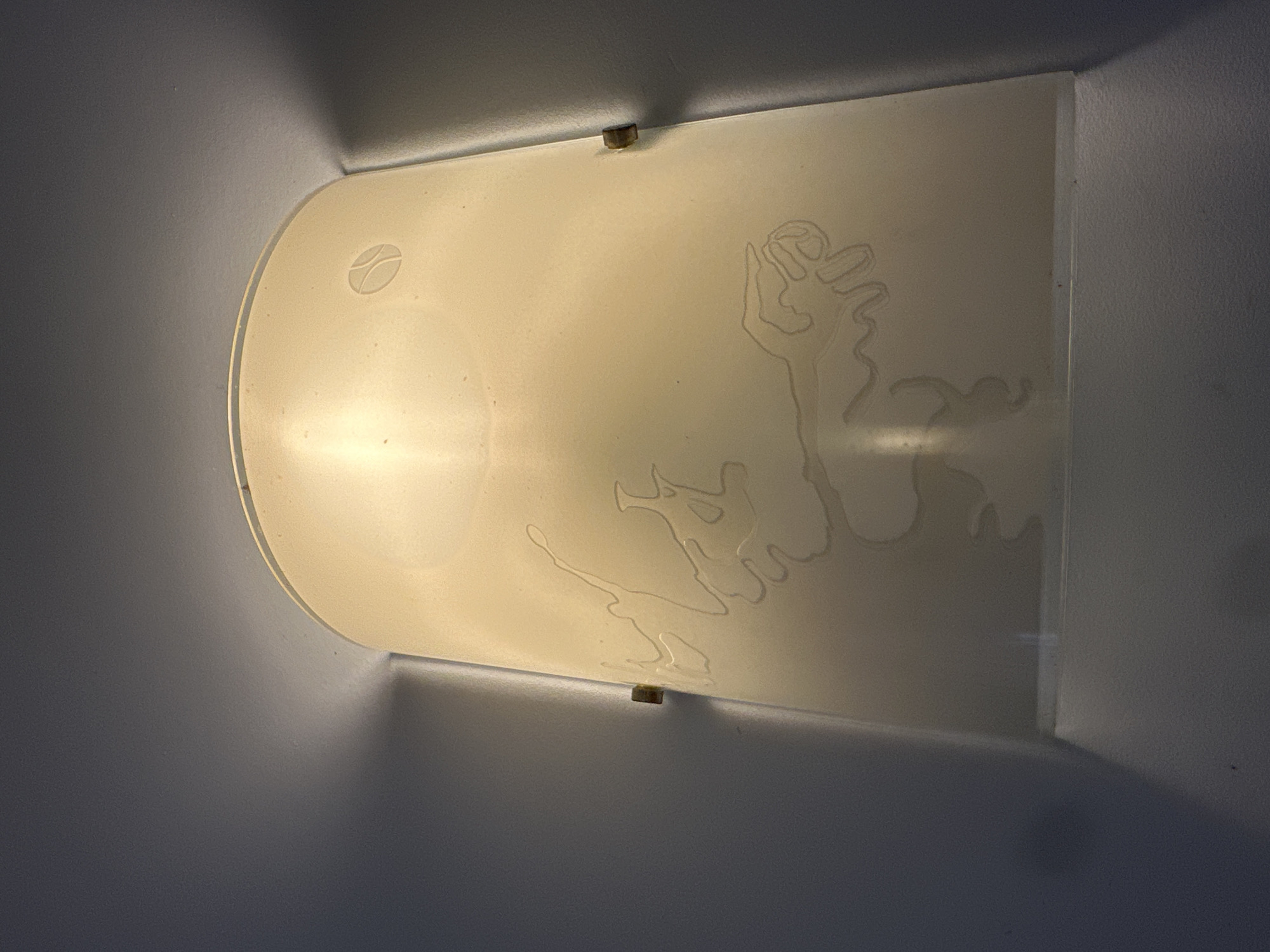 A semi-cylindrical wall sconce with an etched design of abstract figures and a circular emblem at the top is mounted on a light-colored wall. The sconce emits a warm, yellow light, creating shadows on the wall. The etched design appears to be a stylized representation of a creature with multiple limbs or appendages, and the circular emblem is a simple outline of two curved shapes intersecting. The sconce is held in place by small, brass-colored brackets visible on either side. The overall impression is of a decorative light fixture in a dimly lit interior space.