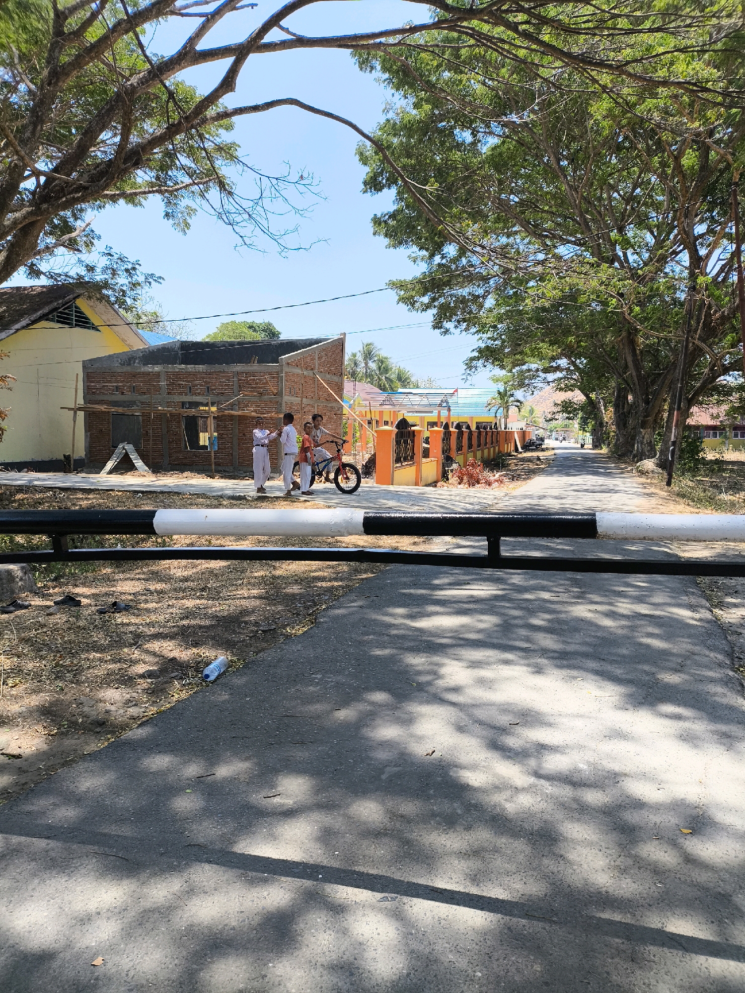 A street scene in Unknown, Indonesia, features a road lined with trees casting dappled shadows on the pavement. In the foreground, a black and white striped barrier stretches across the road. To the left, a building under construction is visible, with exposed brickwork and scaffolding. Three young people, appearing to be students in white uniforms, are gathered near the construction site. One of them is standing next to a red bicycle, with another person sitting on it and two others standing. Further down the road, there are more buildings with colorful roofs and fences. The sky is clear and blue. A discarded plastic water bottle lies on the side of the road.