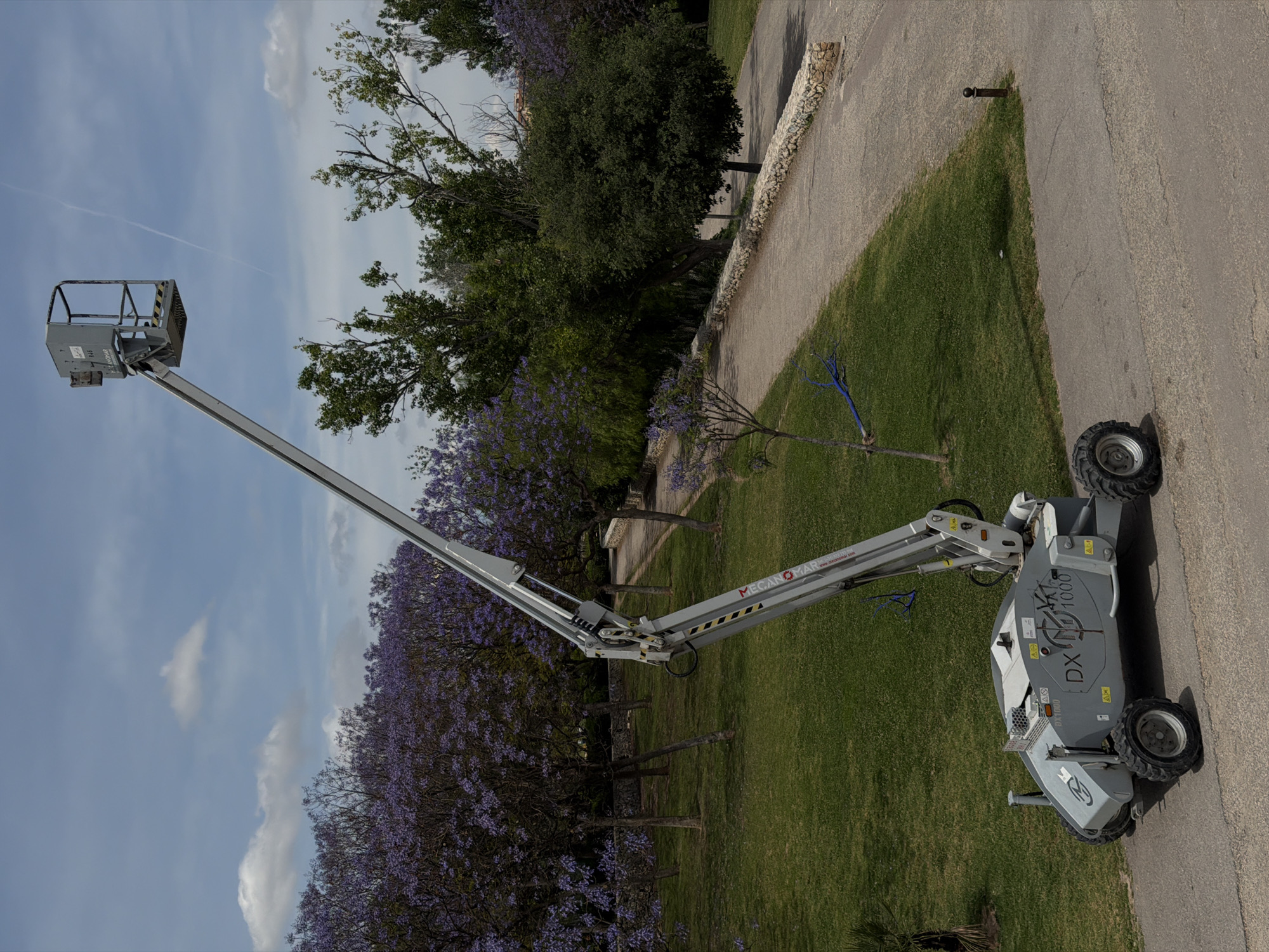 A gray articulated lift is parked on a paved surface beside a grassy area in Valencia, Spain. The lift's bucket is extended upwards towards a blue sky dotted with clouds. Trees with purple flowers are visible on the left side of the image, adding a pop of color to the scene. A paved pathway runs along the right side, bordered by a low stone wall.