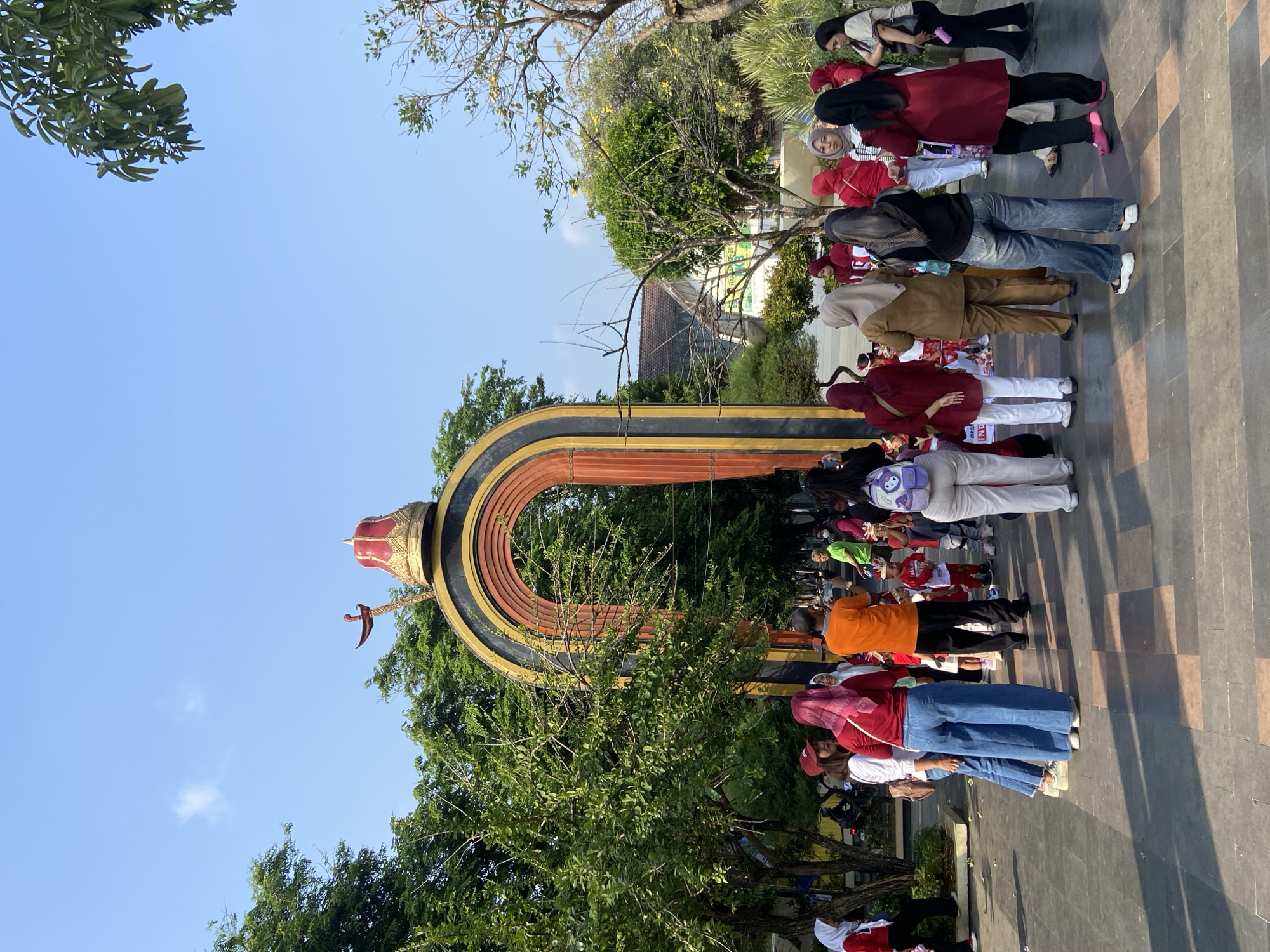 The image depicts an outdoor scene in Kecamatan Pati, Indonesia, featuring a large arched gateway adorned with a red and gold headdress and a ceremonial sword. A crowd of people, primarily women and children, are gathered around the gateway. Many are wearing red and white attire, suggesting a festive occasion or national celebration. The ground is paved with square tiles, and the area is lined with trees, indicating a park or public square. The sky is clear and blue, with some scattered clouds, and the sun is casting shadows on the ground. The people are diverse in age and are engaged in various activities, such as standing, walking, and observing. Some individuals are taking photos with their phones. The atmosphere appears to be lively and communal.